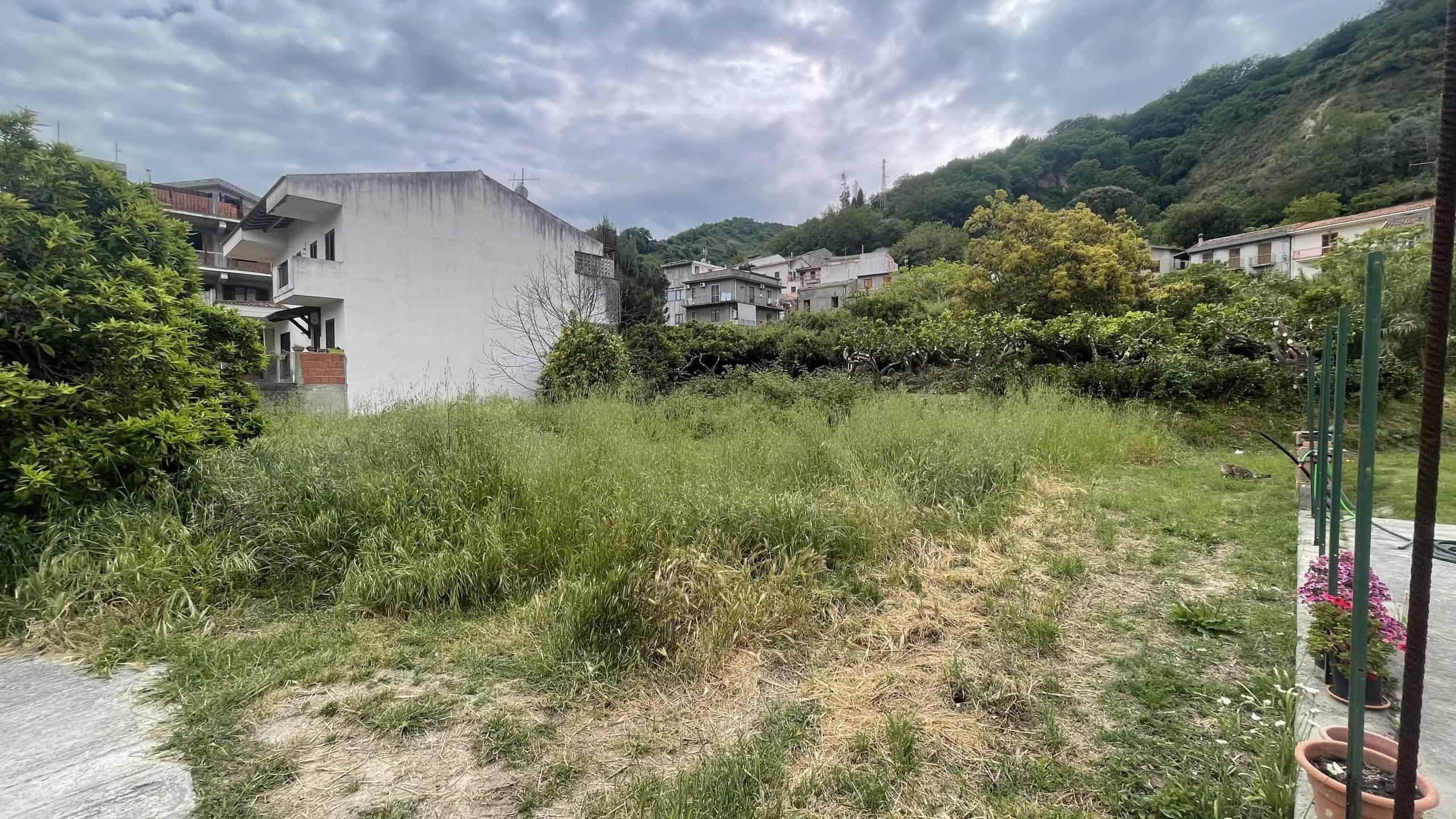 Prato di terreno verde con erba alta e piante, sfondo di case e colline verdi sotto un cielo nuvoloso, ideale per immobili residenziali o progetti di sviluppo immobiliare in zona naturale.