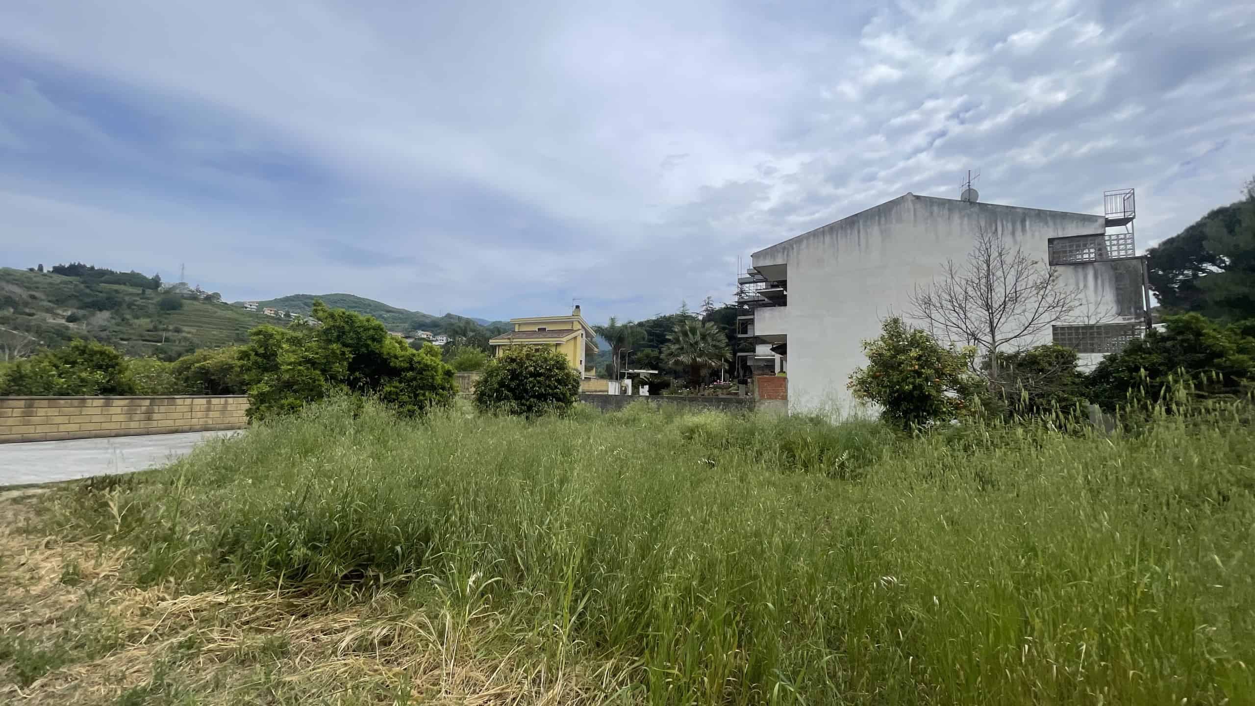 Prato verde e aperto con edifici residenziali e colline sullo sfondo, cielo nuvoloso, natura e tranquillità.