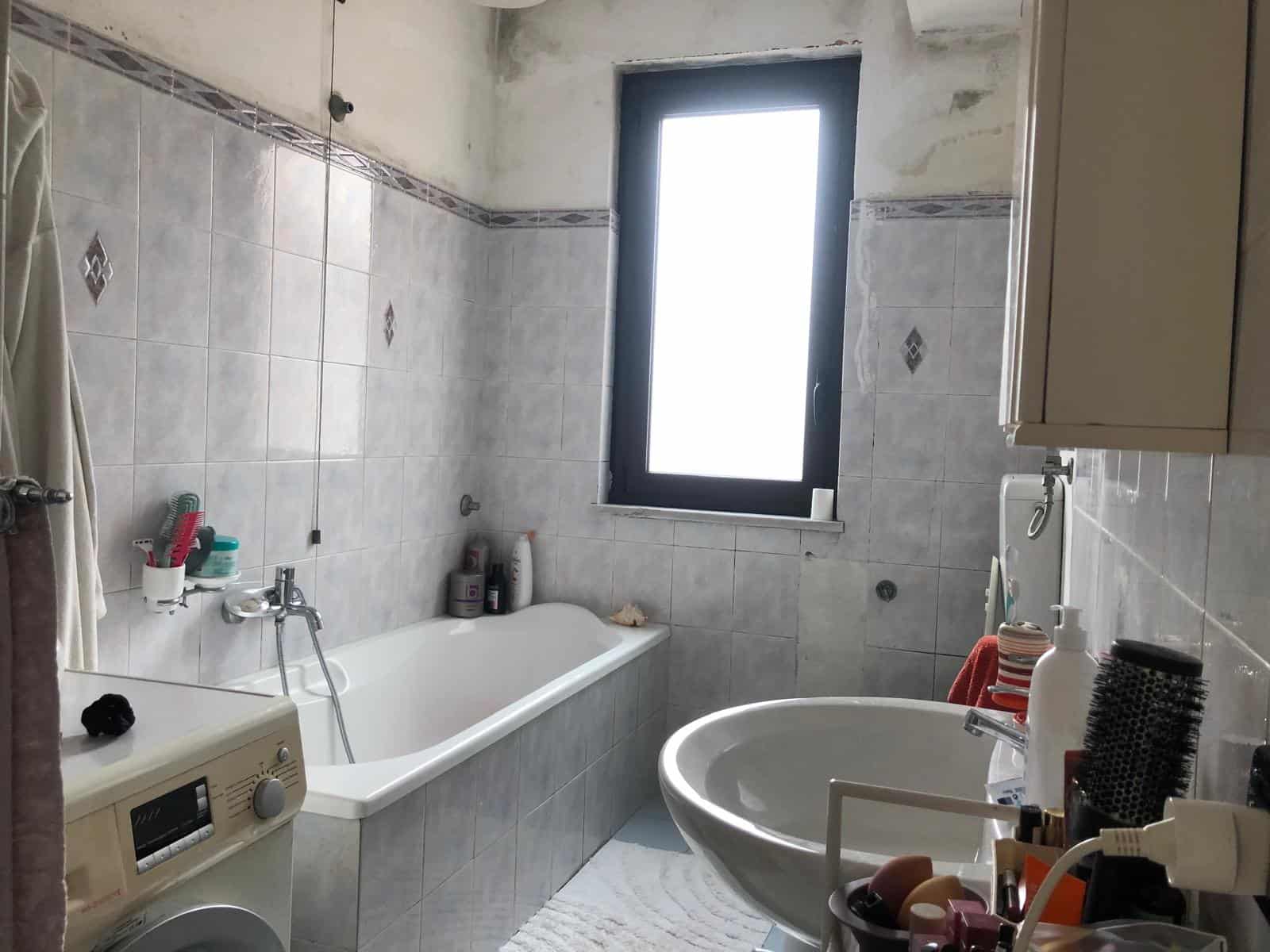 Lavabo e vasca da bagno in un bagno moderno con pareti rivestite in piastrelle grigie, finestra grande che permette l'ingresso di luce naturale, e vari accessori per la cura personale e la pulizia.