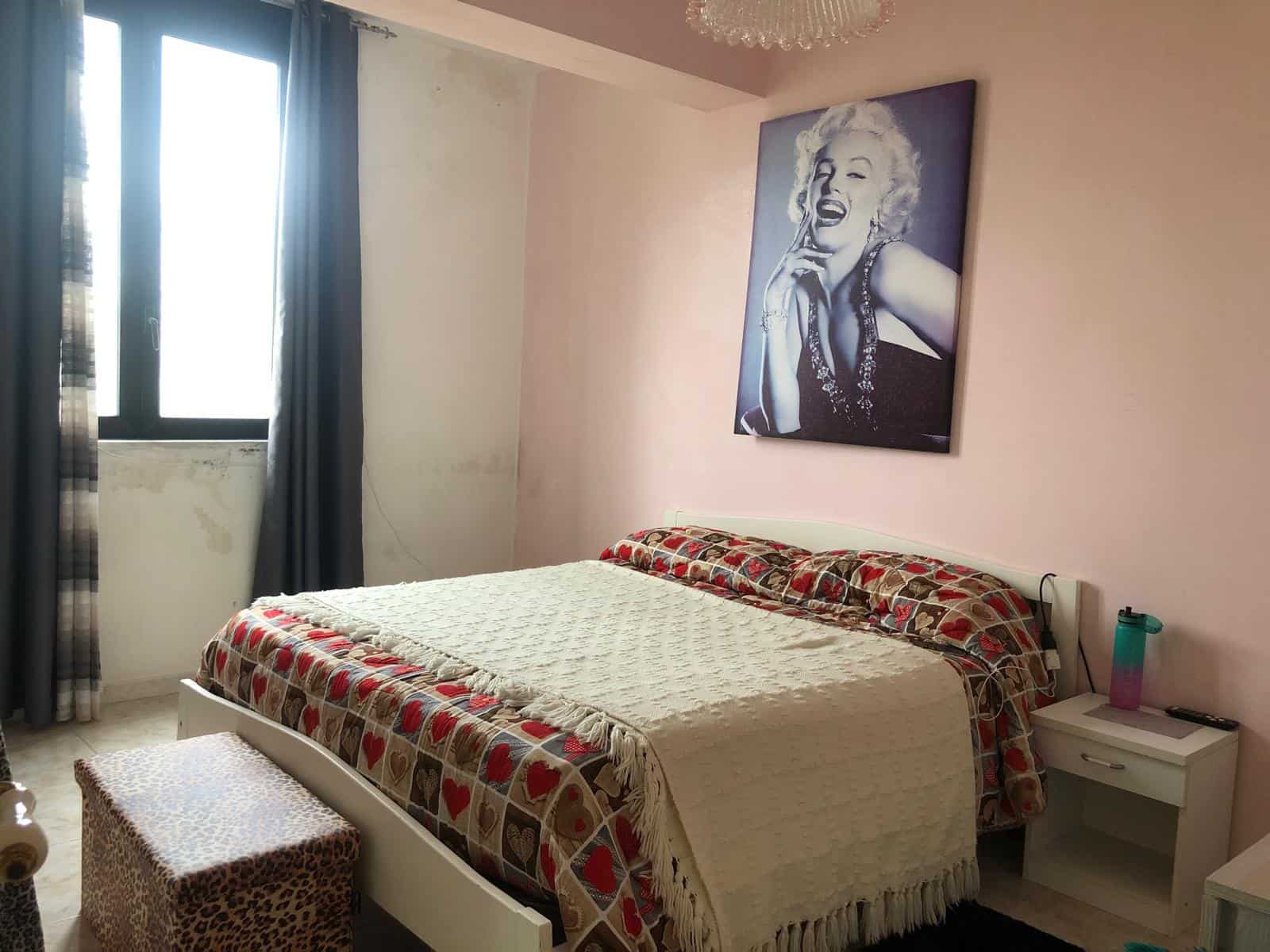 Letto matrimoniale con copriletto decorativo, in una camera da letto luminosa con pareti rosa e un quadro di Marilyn Monroe appeso alla parete. Finestra con tende scure permette l'ingresso di luce nat.