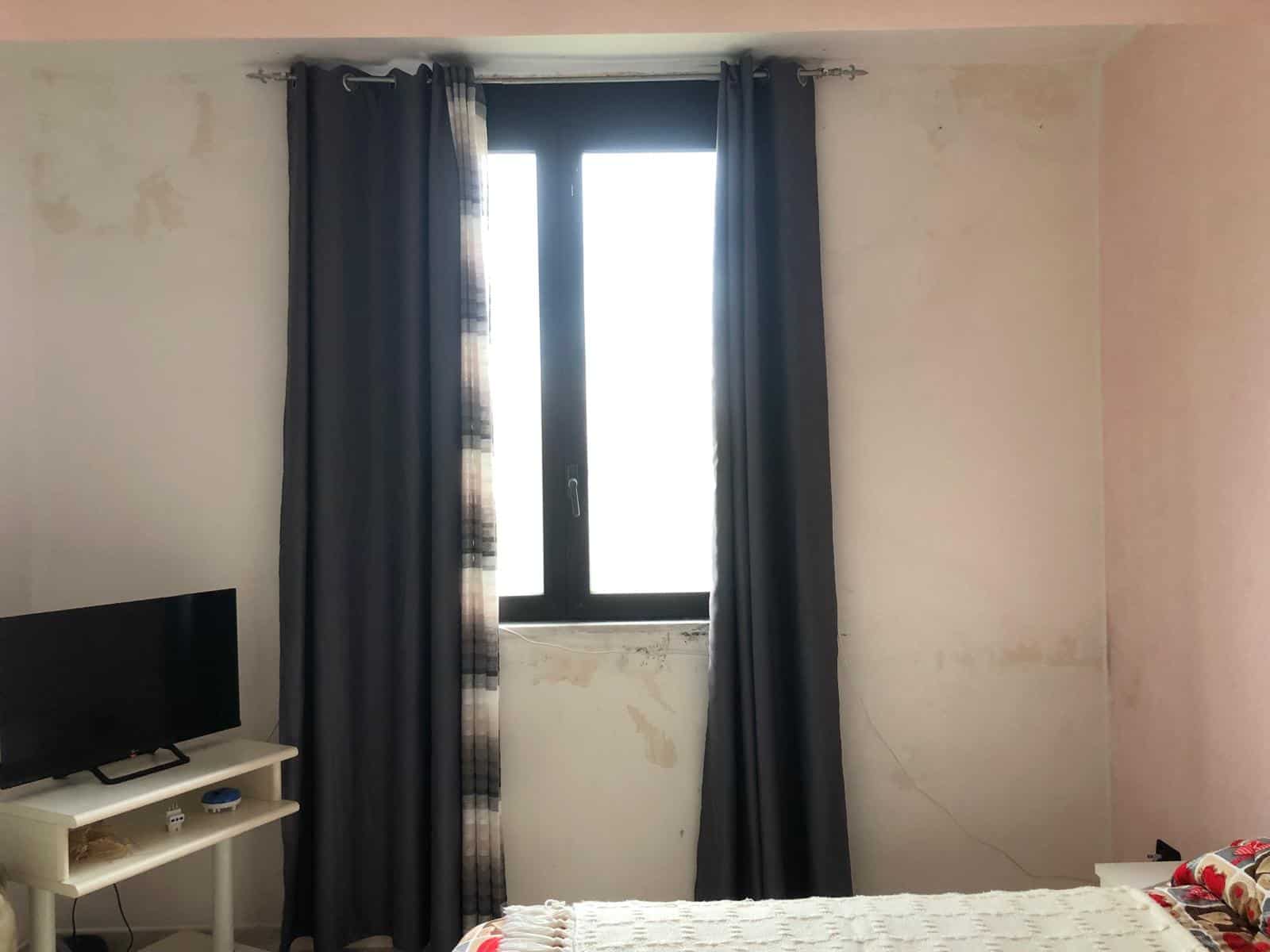 Tende scure nere davanti a una finestra in una stanza con pareti bianche e un letto con biancheria colorata, ambiente luminoso e accogliente.