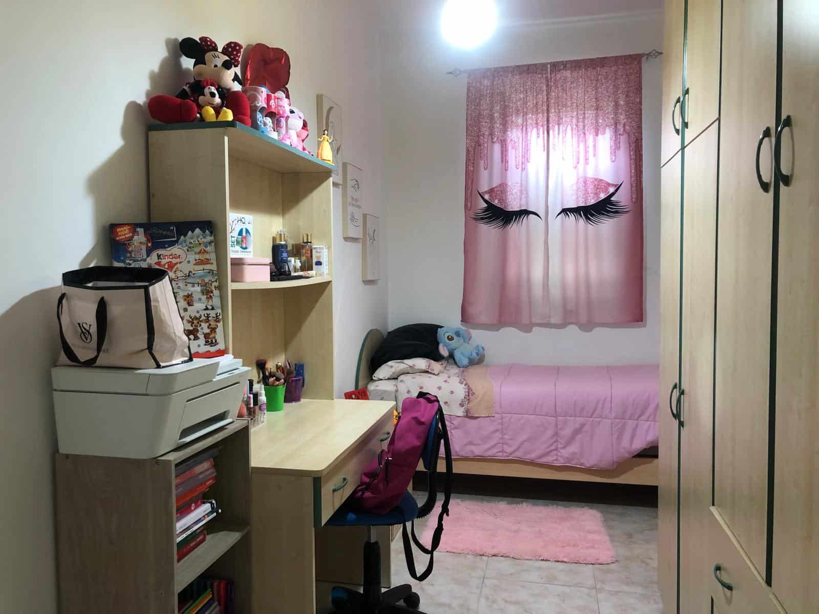 Le immagini mostrano una camera da letto per bambini con decorazioni femminili, tra cui tende con ciglia nere e un letto con cuscini e peluche, mobili in legno chiaro e scaffali pieni di giocattoli e.