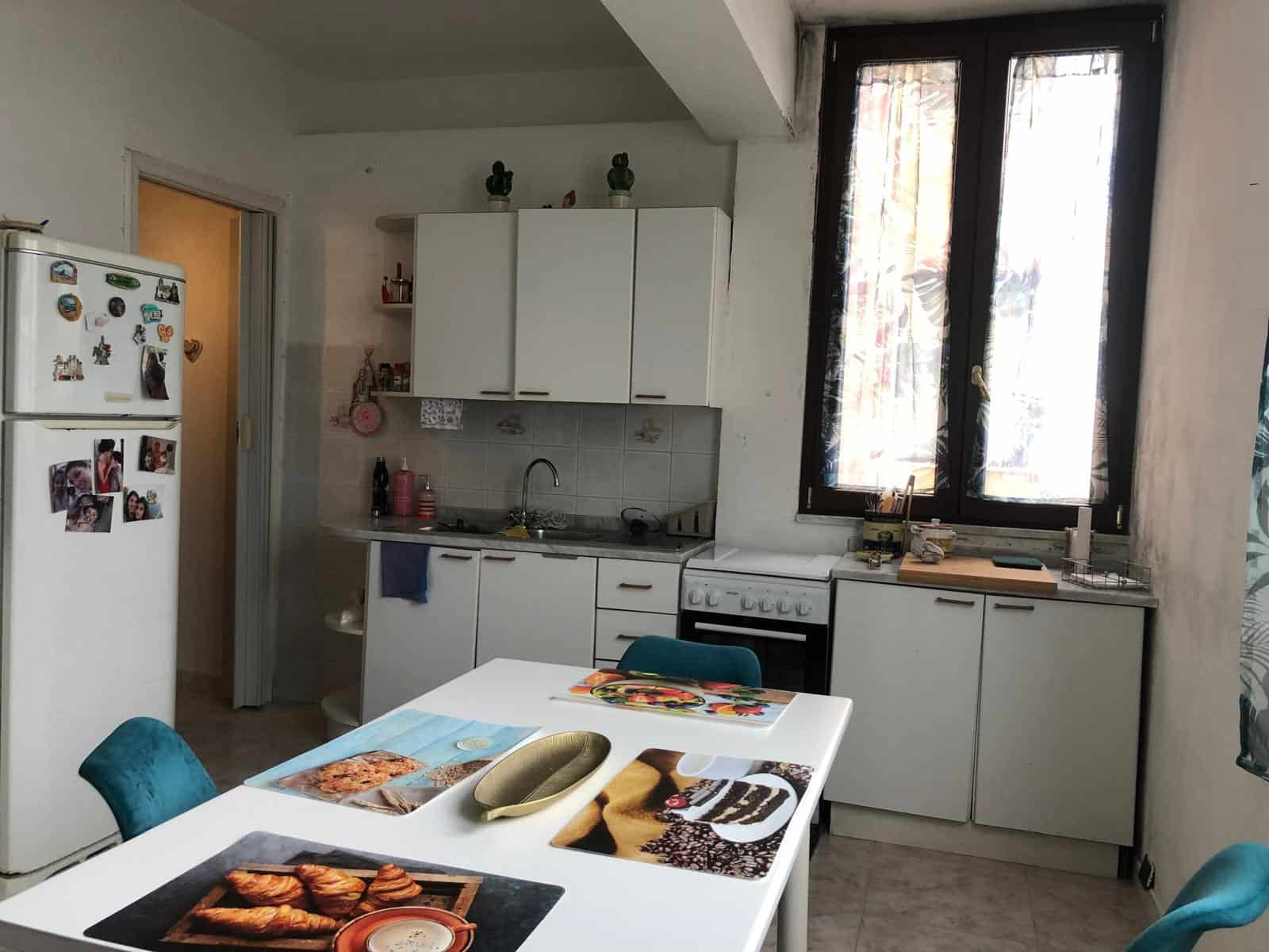 Porte aperte su una cucina moderna e luminosa con mobili bianchi, tavolo da pranzo con piatti e decorazioni, e grandi finestre che lasciano entrare molta luce naturale.