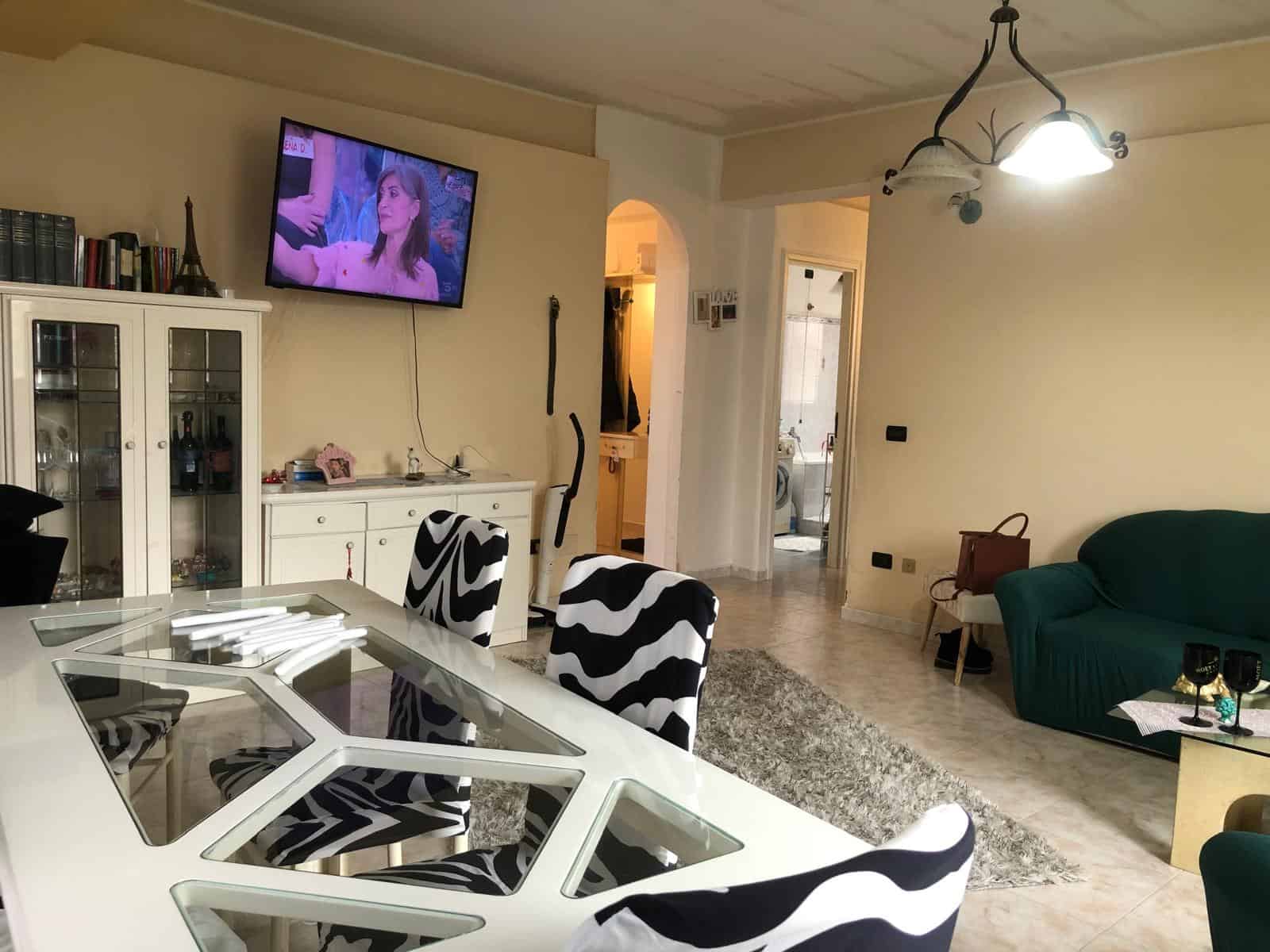Ampia zona living moderna con divano verde, tavolo da pranzo con sedie a righe nere e bianche, televisore a parete e arredi eleganti in un ambiente luminoso e accogliente.