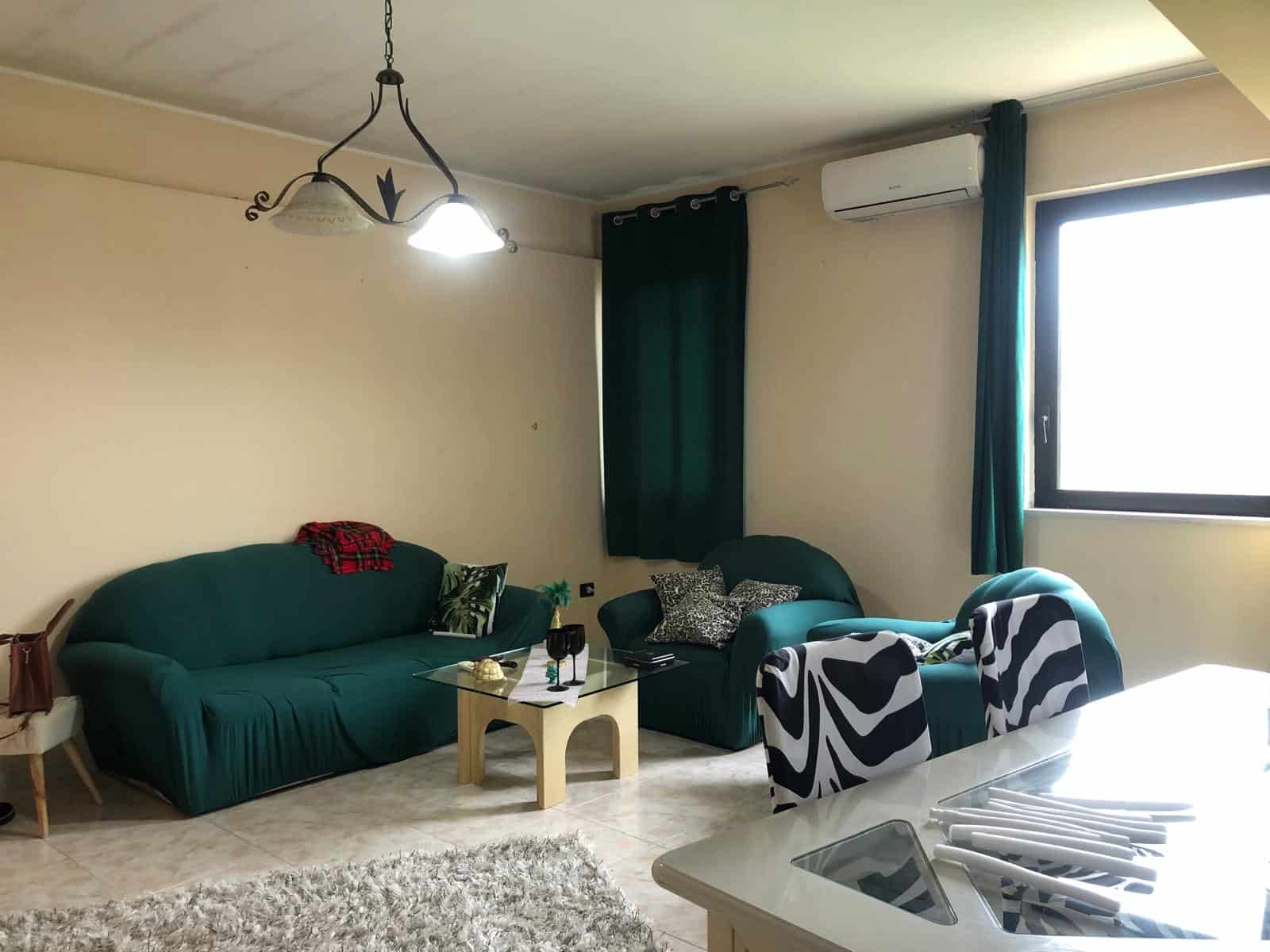 Divano verde in soggiorno con tavolino e sedie con rivestimento a motivi astratti, ambiente luminoso con finestra e aria condizionata, arredamento moderno e accogliente.