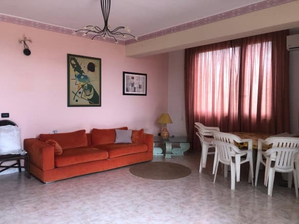 Ampia zona living con divano rosso, tavolo da pranzo con sedie bianche, tende rosa e decorazioni moderne, ideale per soggiorni confortevoli e conviviali.