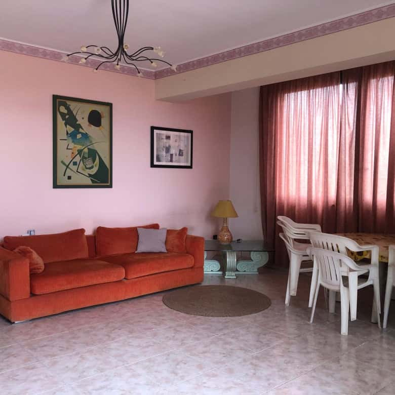 Ampia zona living con divano rosso, tavolo da pranzo con sedie bianche, tende rosa e decorazioni moderne, ideale per soggiorni confortevoli e conviviali.