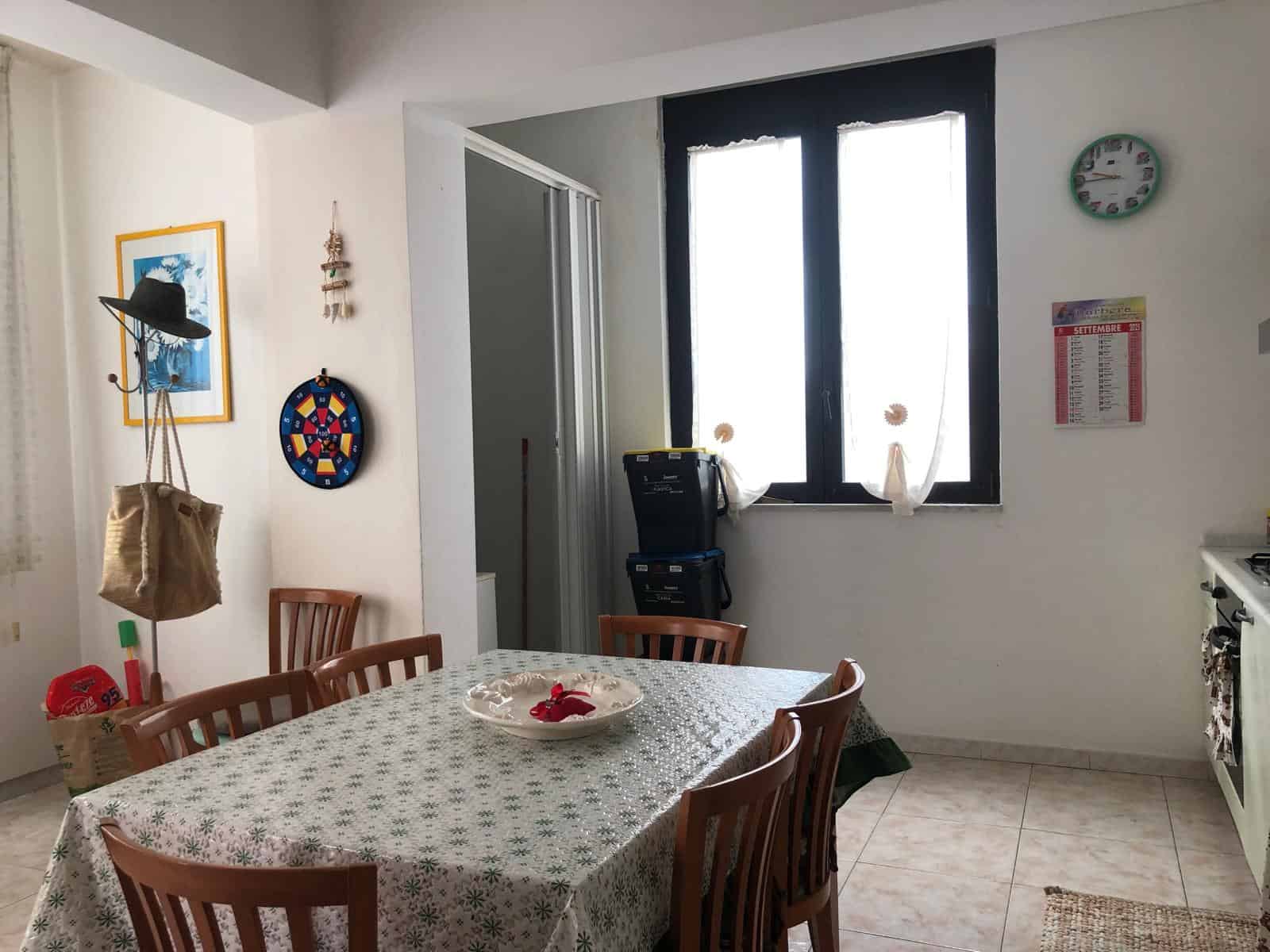 Sedia in legno attorno a tavolo da pranzo con tovaglia floreale in una cucina luminosa e accogliente, finestra grande con tende nere, decorazioni colorate alle pareti, ambiente domestico caldo e invit.