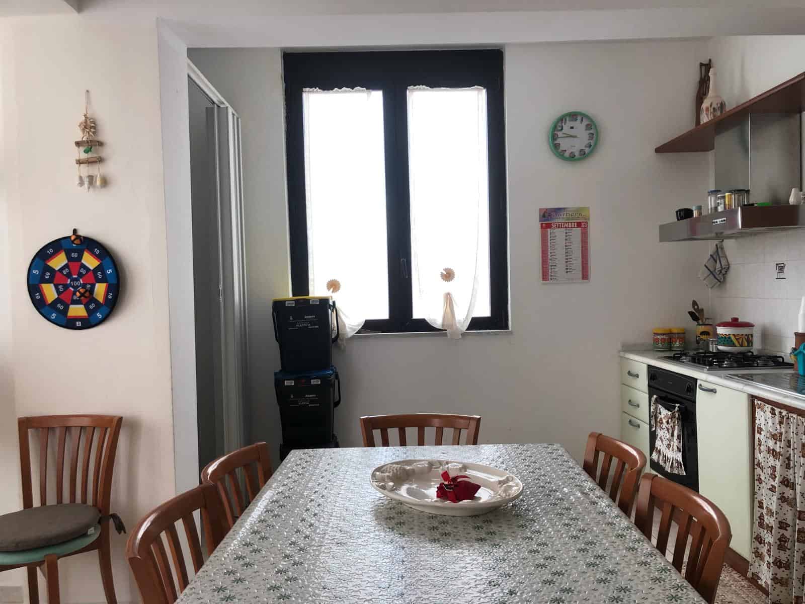 Ampia sala da pranzo luminosa con tavolo decorato, sedie in legno e cucina funzionale, ideale per vivere momenti conviviali in un ambiente accogliente e ben illuminato.