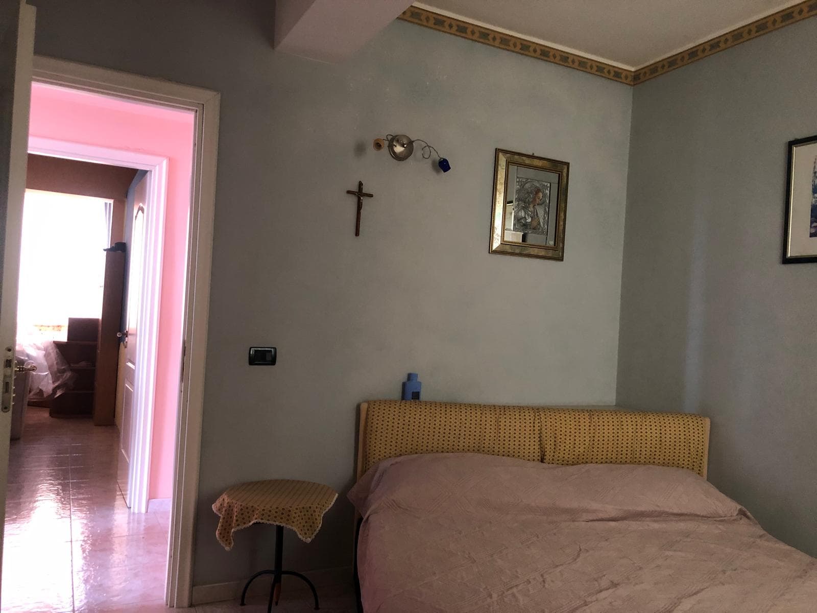 Una stanza da letto luminosa con pareti azzurre, decorazioni religiose e un letto con testiera imbottita color senape, in un appartamento in stile classico.