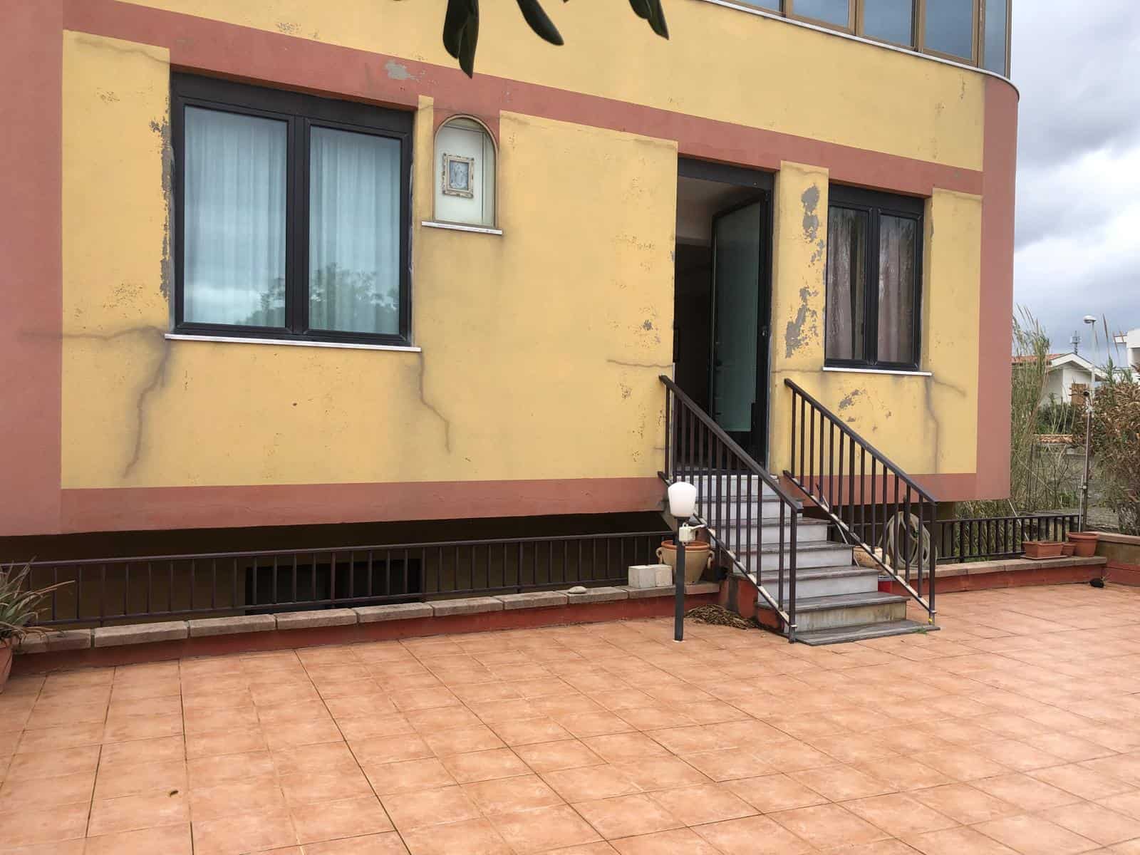 Ampia terrazza esterna con accesso alla casa, pavimentata in piastrelle di ceramica, con vista sulla facciata gialla con finestre e porta d'ingresso.