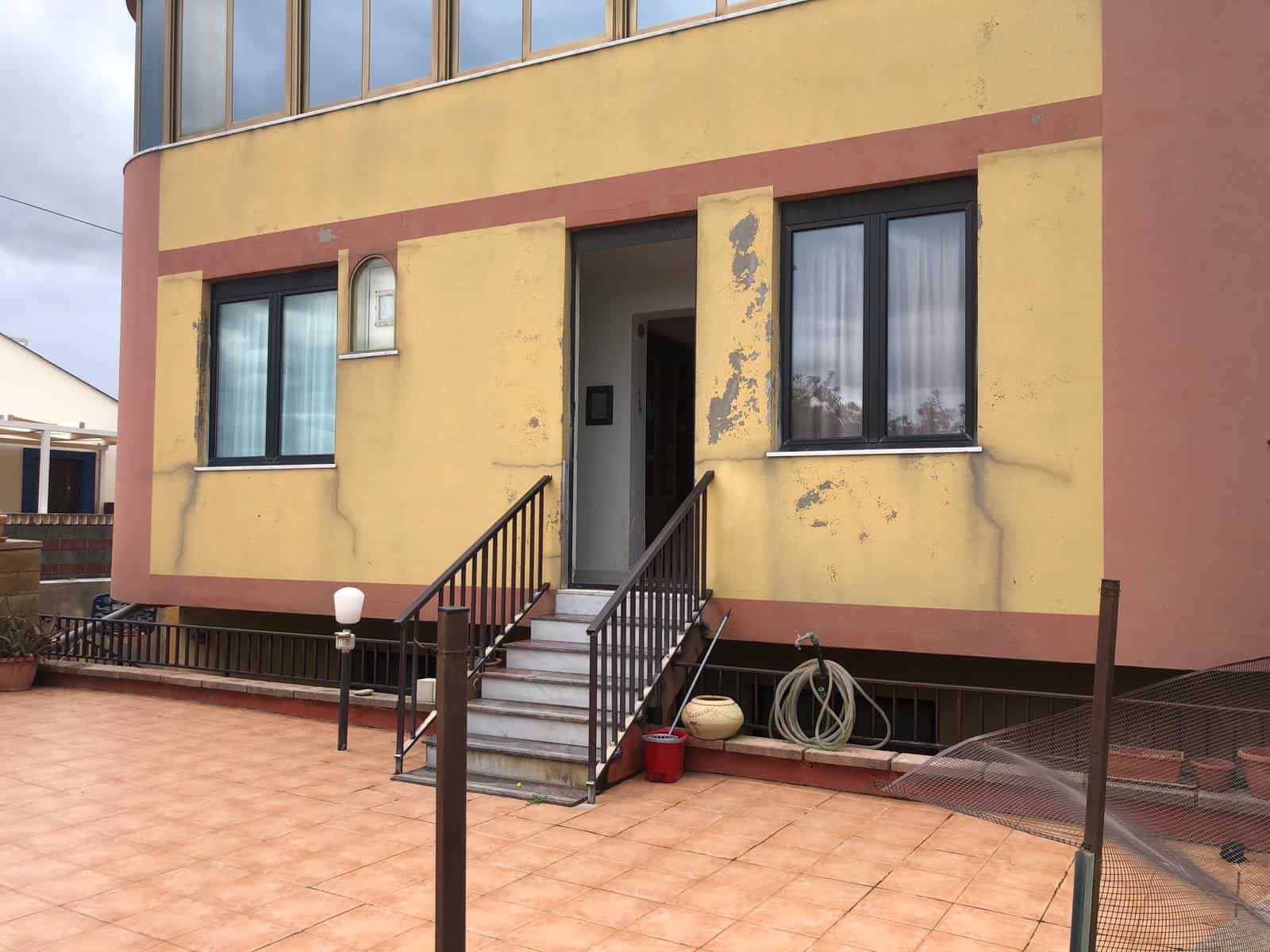 Pannello frontale di una casa con pareti gialle e dettagli in rosso, ingresso con scale e ringhiera in metallo, area esterna pavimentata con accessori e attrezzi, ambiente residenziale.