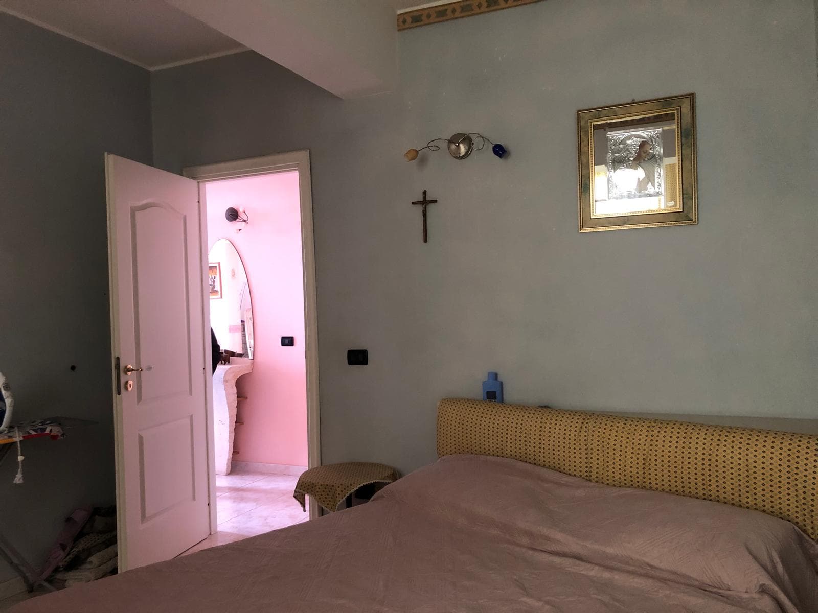 Letto matrimoniale con testiera imbottita in tessuto beige, in una camera da letto luminosa e accogliente con pareti azzurro chiaro, decorazioni religiose e un quadro sopra il letto.