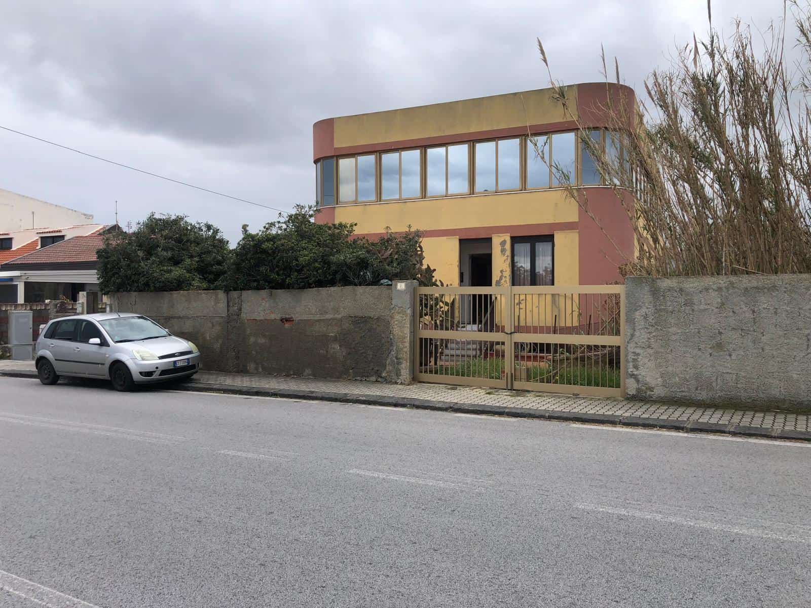 Barriera di casa moderna con pareti in cemento e recinzione in metallo, situata lungo una strada asfaltata con un'auto parcheggiata davanti. Edificio residenziale con grandi finestre e un design archi.