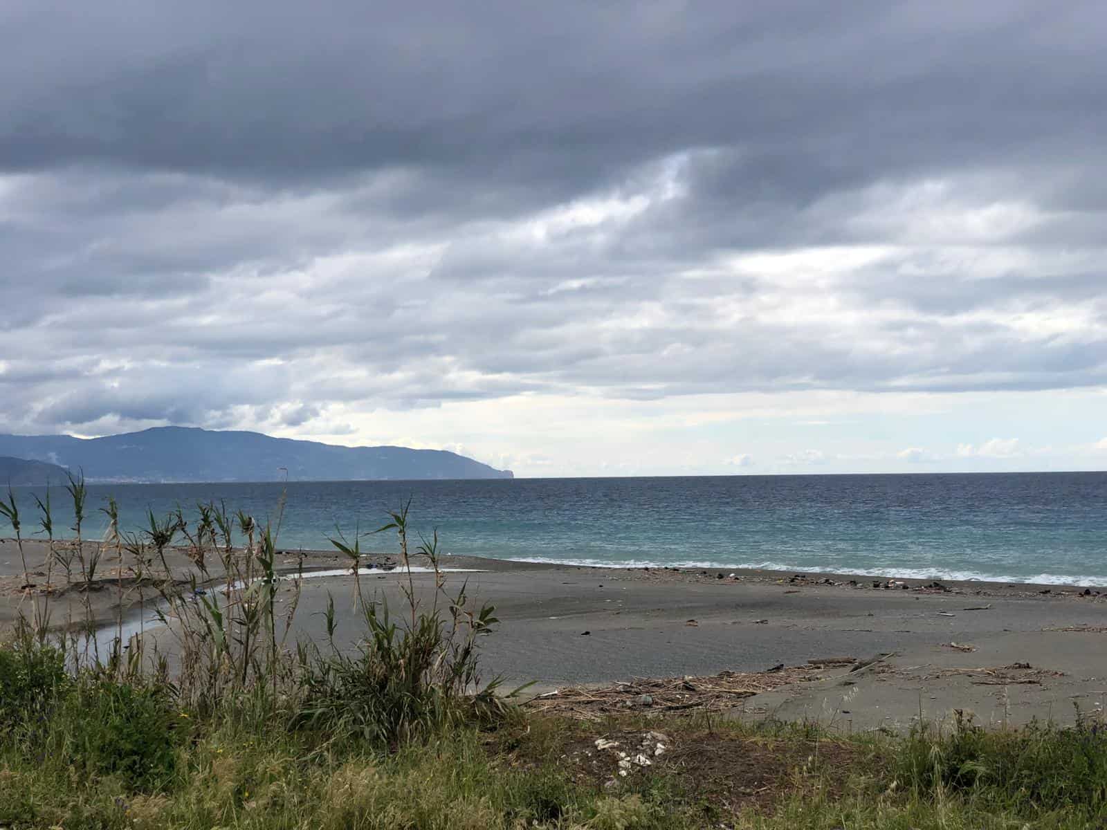 Spiaggia con sabbia grigia e vegetazione, mare calmo sotto un cielo nuvoloso, con montagne sullo sfondo, atmosfera tranquilla e naturale, ideale per relax e passeggiate sulla costa.