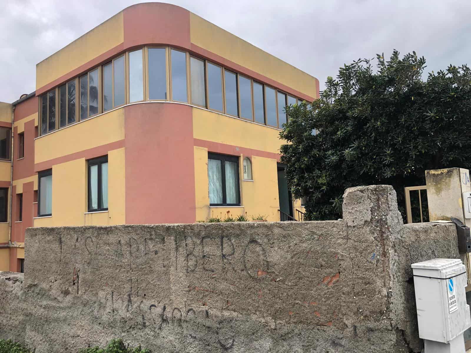 Cancello in cemento con scritte e casetta postale, edificio residenziale moderno con grandi finestre e pareti colorate in tonalità di giallo e rosa, circondato da alberi e vegetazione.