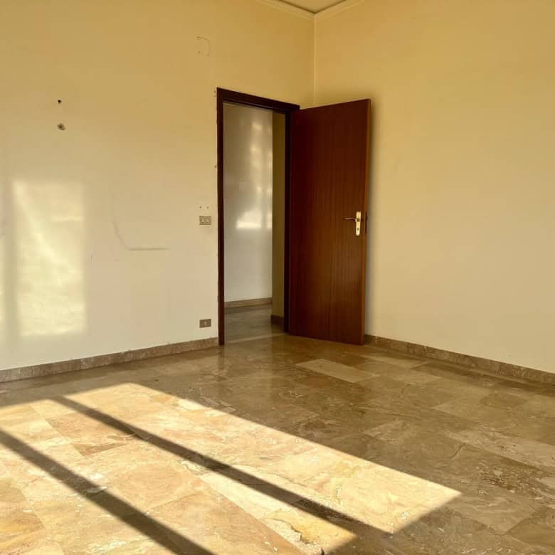 Stanza vuota con pareti beige, pavimento in marmo e porta aperta che lascia entrare luce naturale.