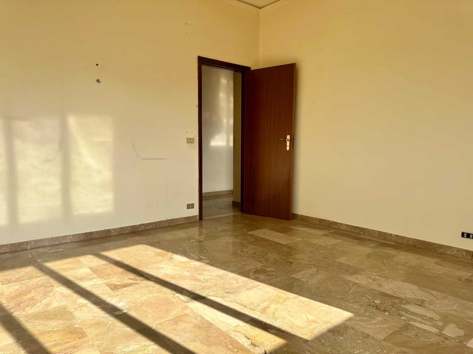 Stanza vuota con pareti beige, pavimento in marmo e porta aperta che lascia entrare luce naturale.