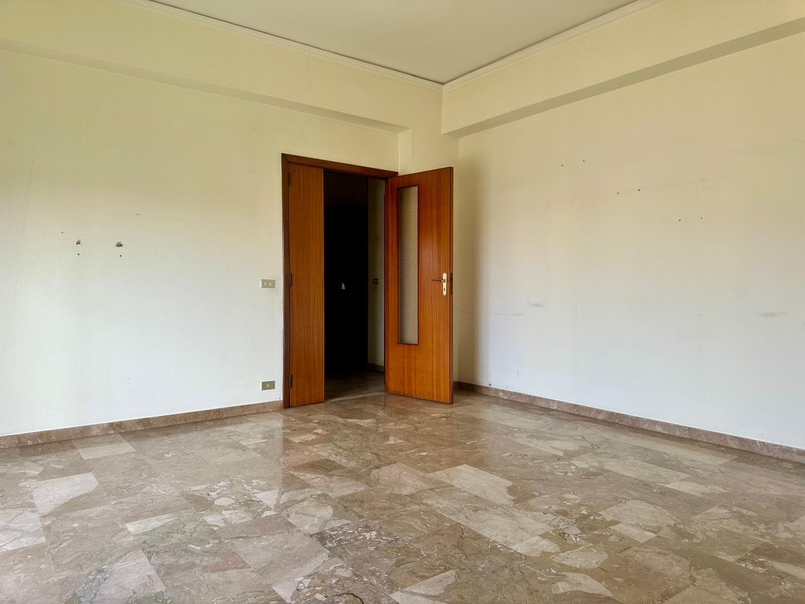 Ampia stanza vuota con pareti bianche e pavimento in marmo, porta in legno aperta che conduce a un'altra stanza, ambiente luminoso e spazioso, ideale per arredamento e personalizzazione.