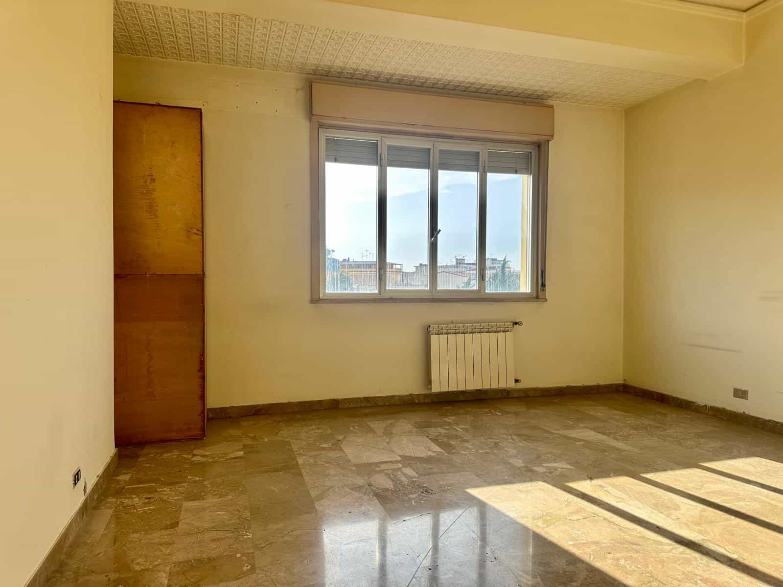 Ampio soggiorno luminoso con grande finestra che offre vista panoramica sulla città, pavimento in marmo e pareti neutre, ideale per arredamento moderno e accogliente.