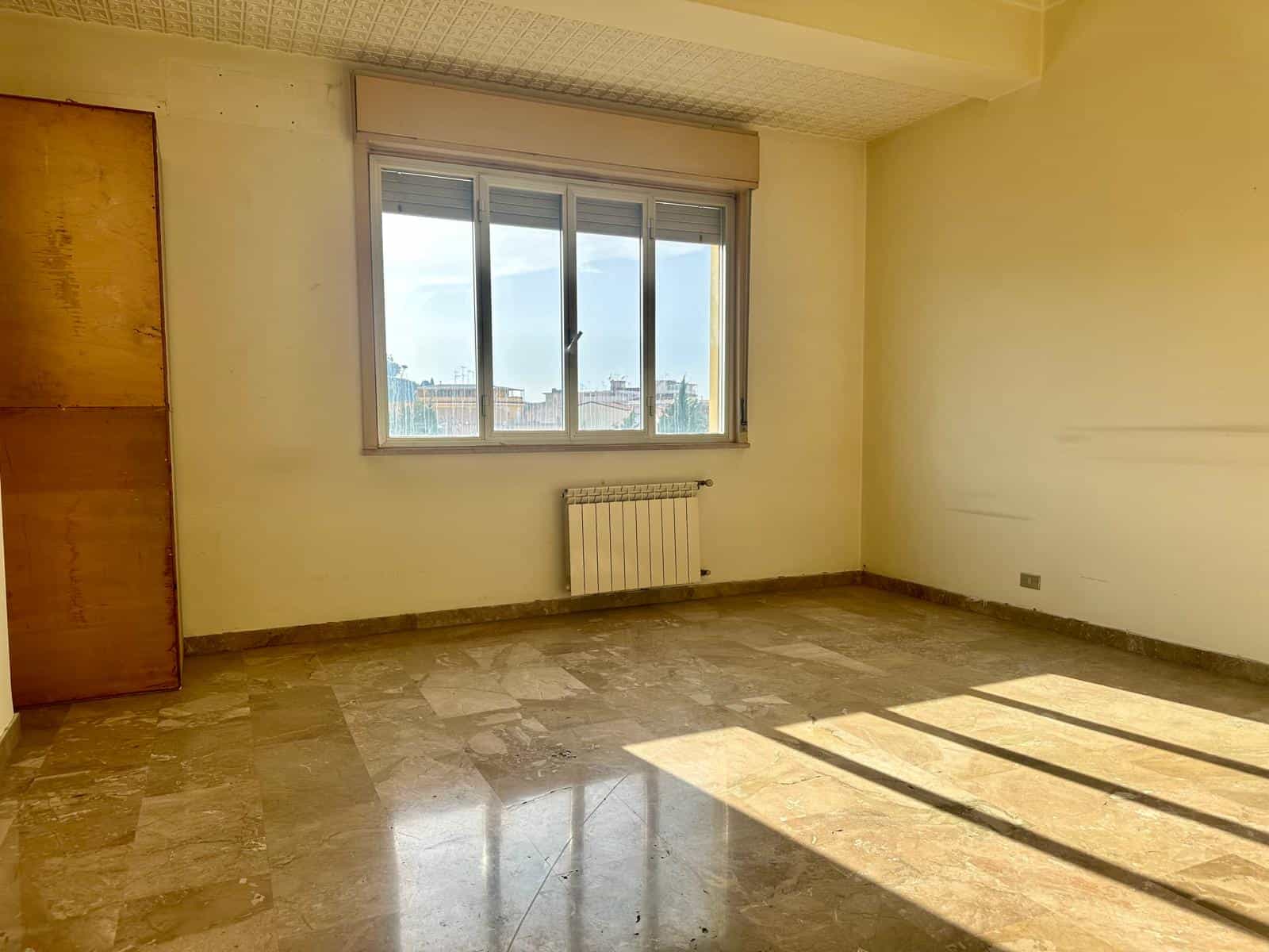 Stanza vuota con grande finestra e vista sulla città, pavimento in marmo e pareti beige, ambiente luminoso e spazioso.