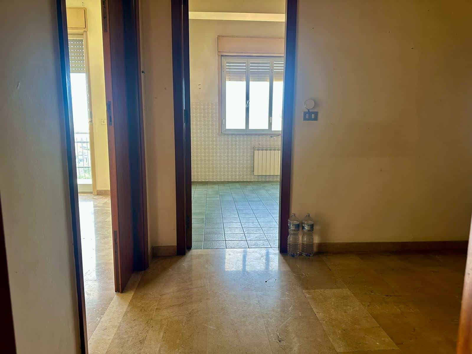 Interno di un appartamento vuoto con pareti beige, pavimento in piastrelle e finestra che lascia entrare luce naturale.