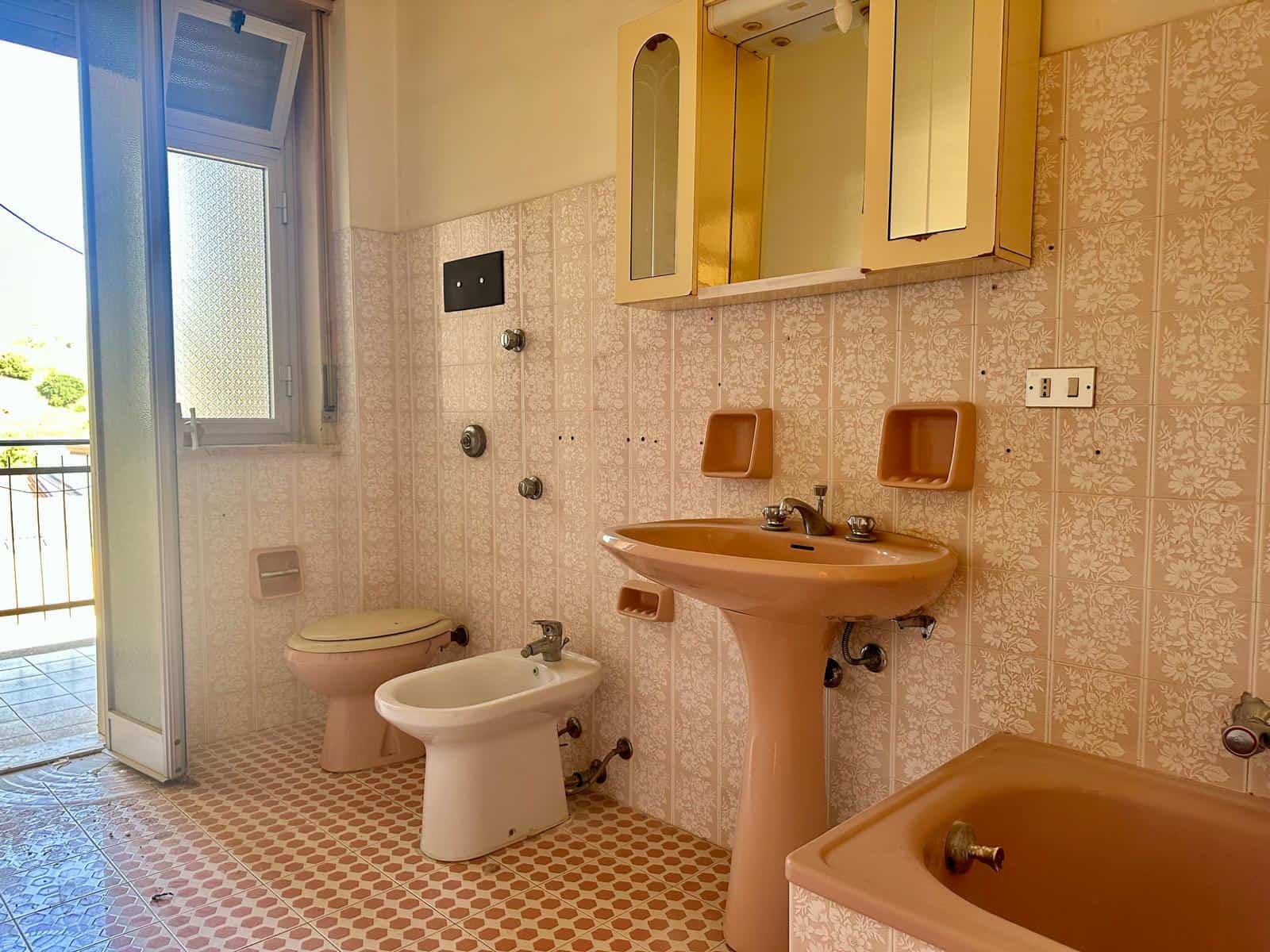 Lavabo e servizi igienici in bagno con pareti rivestite di piastrelle beige e pavimento a mosaico. Finestra aperta che permette l'ingresso di luce naturale e vista sul balcone esterno. Arredo bagno in.