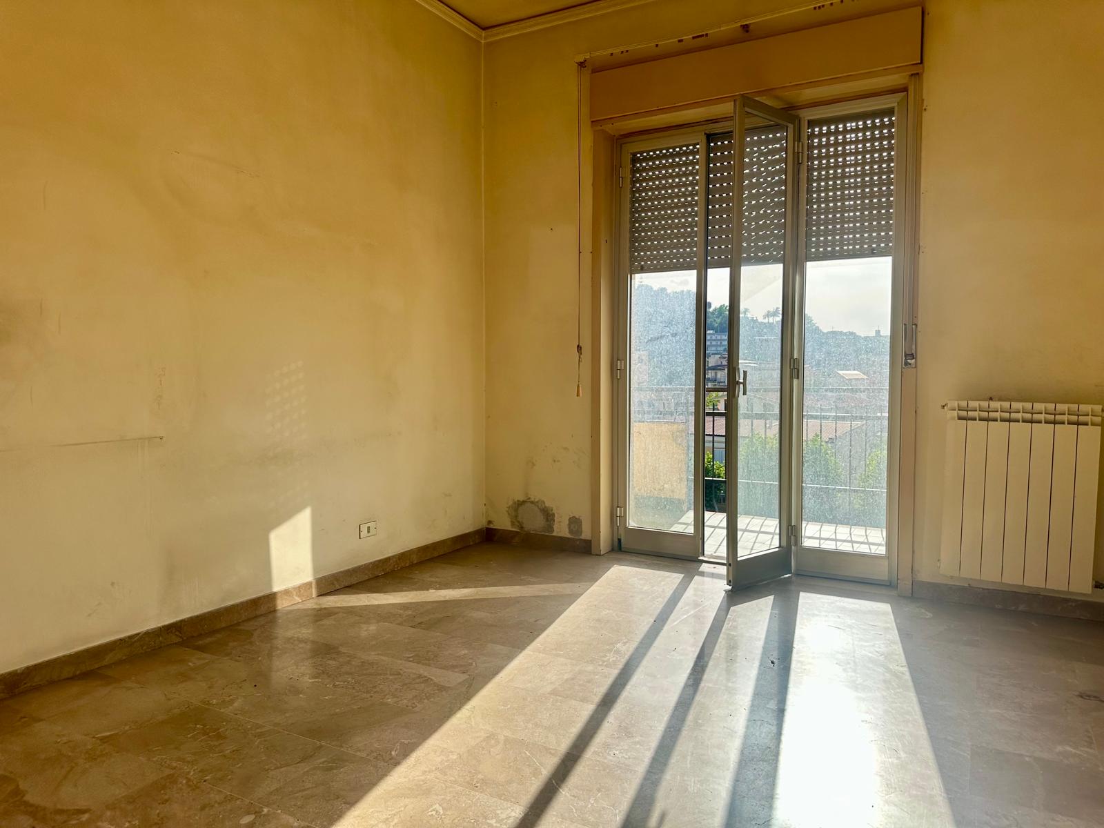 Porte finestra con balcone e vista panoramica su un appartamento in vendita o affitto, illuminato dalla luce naturale del sole, con pareti neutre e pavimento in marmo.