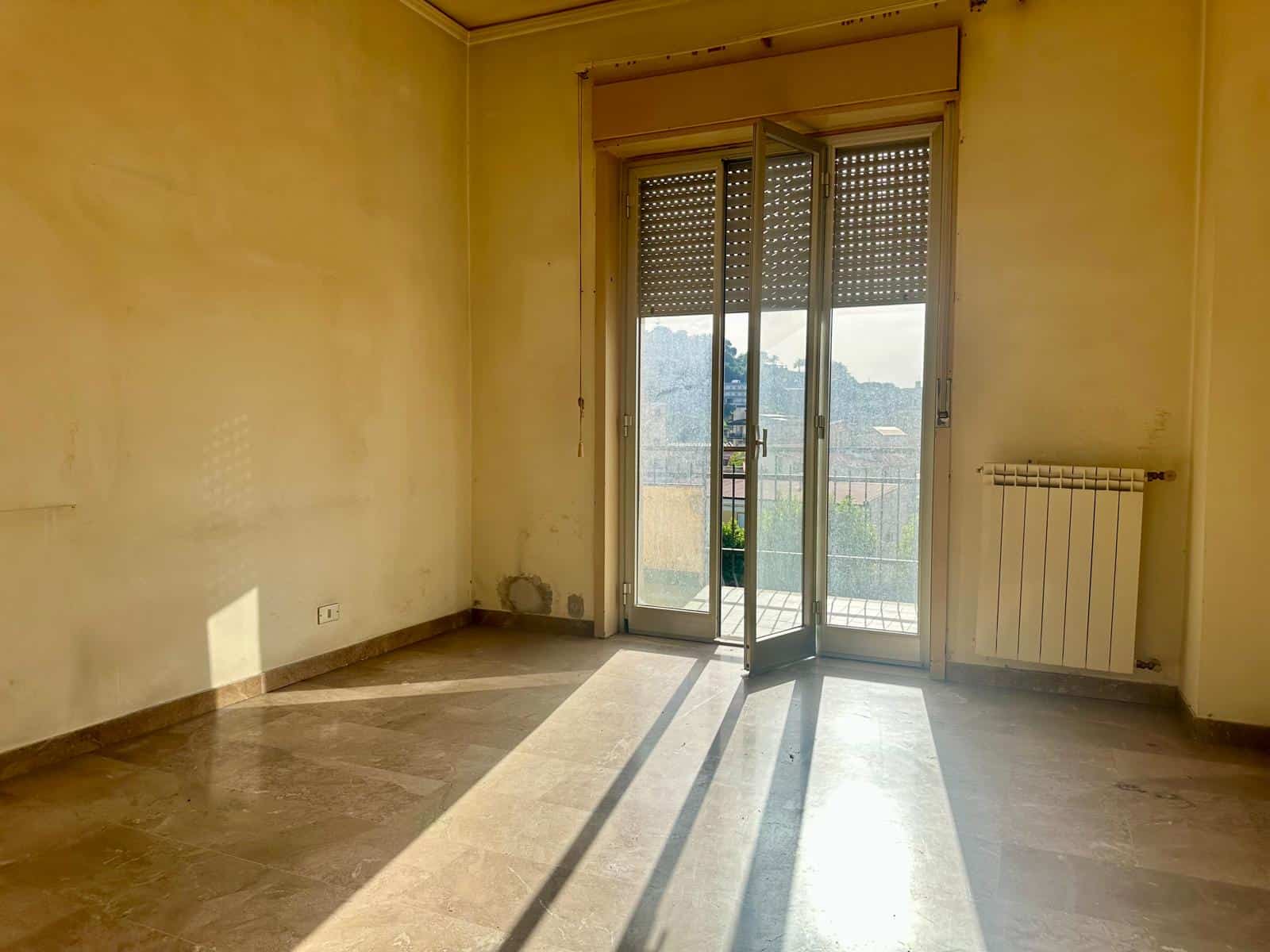 Stanza vuota con pareti gialle, pavimento in marmo e porta finestra che apre su un balcone, illuminata dalla luce naturale del sole.