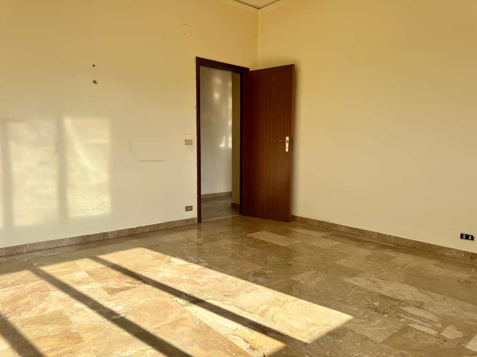 Una stanza vuota con pareti beige e pavimento in marmo, illuminata dalla luce naturale che entra da una finestra non visibile, con una porta aperta che conduce a un'altra stanza.