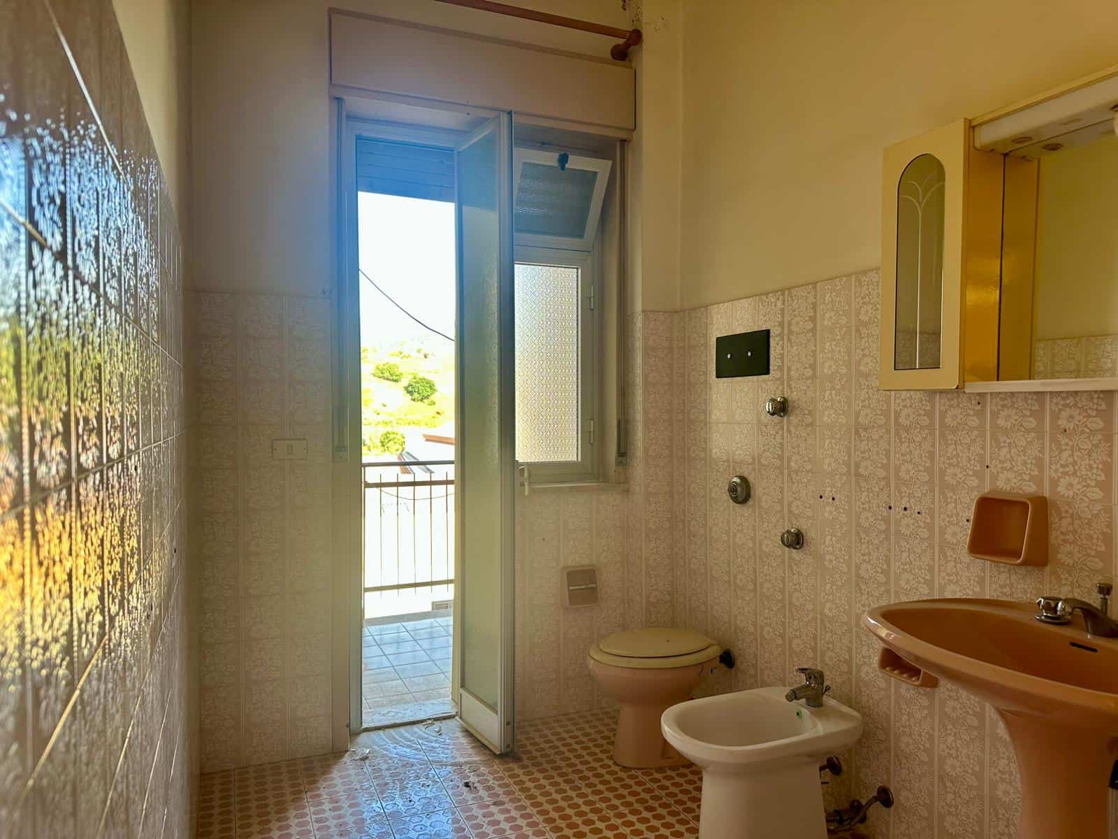 Bagno con accesso a un balcone, dotato di sanitari e lavabo in stile vintage, illuminato dalla luce naturale proveniente dalla finestra aperta, con vista sul balcone e l'esterno.