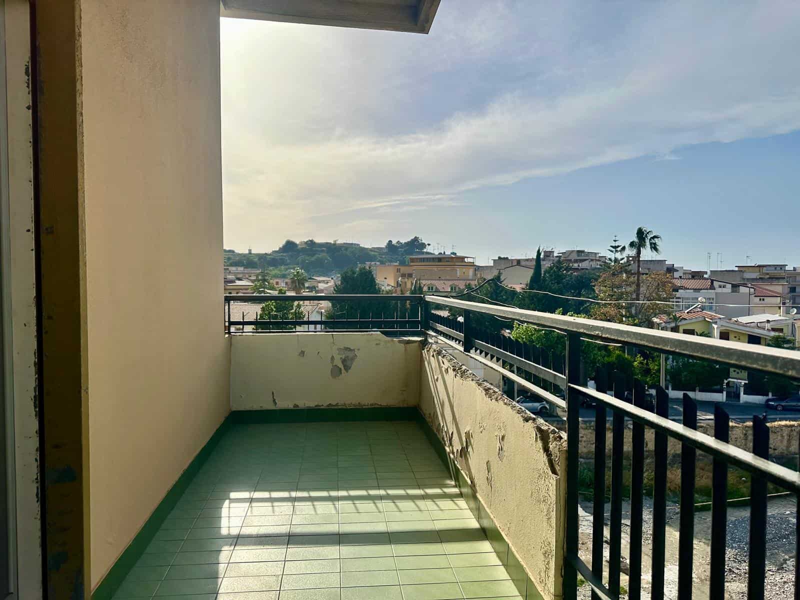 Balcone di un appartamento con vista panoramica sulla città e cielo sereno, pavimento in piastrelle verdi e ringhiera in metallo nero, ambiente luminoso e spazioso.