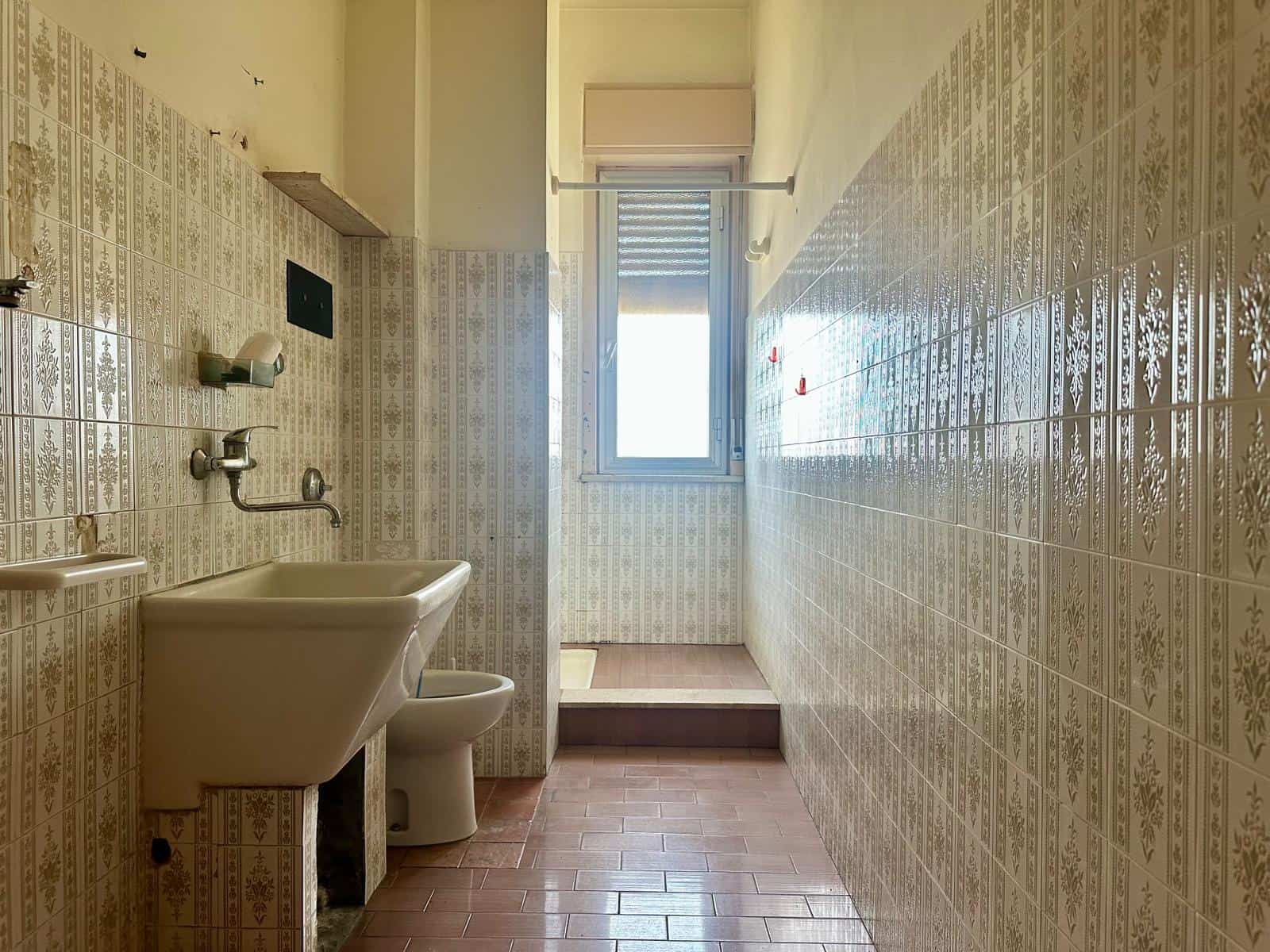 Lavabo e servizi igienici in un bagno con pareti rivestite di piastrelle decorative e finestra luminosa, ideale per annunci immobiliari e ristrutturazioni.