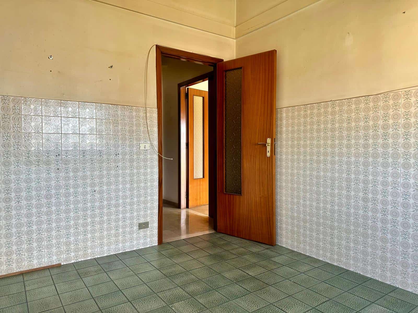 Aria di una stanza vuota con pareti rivestite in piastrelle decorative e pavimento in piastrelle verdi, porta in legno aperta che conduce a un'altra stanza luminosa.