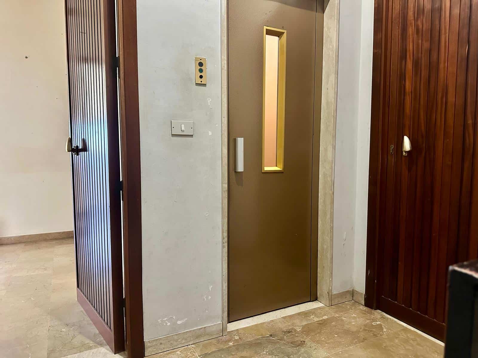 Ascensore moderno in edificio residenziale con pareti in metallo color oro e finestra verticale, situato tra pareti in legno e pareti in cemento chiaro, pavimento in pietra naturale.