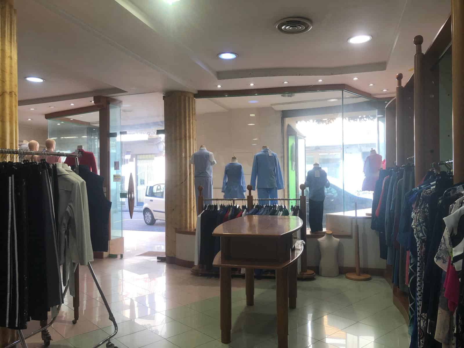 Cattura di un negozio di abbigliamento con manichini e vestiti esposti, ingresso con vetrina e clienti che fanno shopping, ambiente luminoso e accogliente.