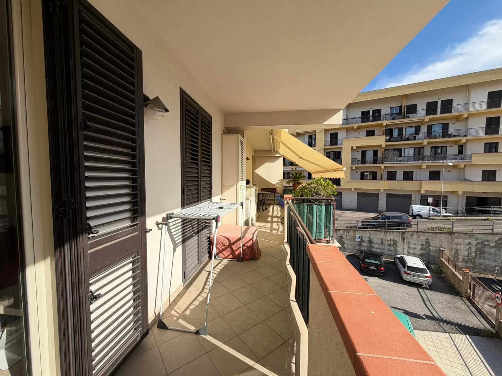 Balcone di un appartamento con vista sulla strada e su edifici residenziali, con arredi da esterno e tende parasole, in un complesso residenziale moderno.