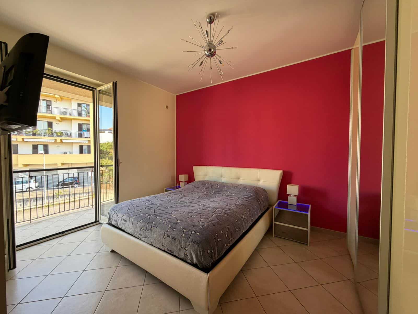 Letto matrimoniale in camera da letto con parete rosa, finestra con vista su balcone e mobili moderni, ambiente luminoso e accogliente.