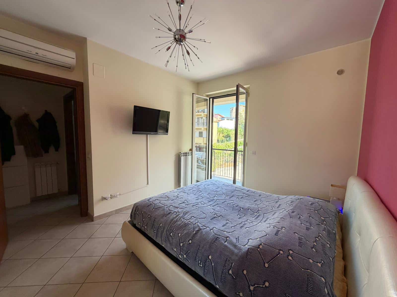 Letto matrimoniale in una stanza luminosa con pareti beige e una parete rosa, finestra aperta che permette l'ingresso di luce naturale, TV a parete e aria condizionata, ambiente accogliente e moderno.