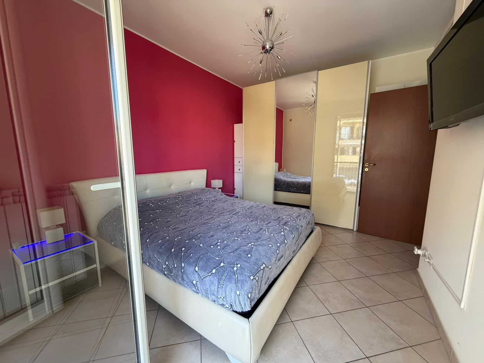 Riproduzione di una camera da letto moderna con pareti colorate, letto matrimoniale con biancheria blu, armadio a specchio e televisore a parete, ambiente luminoso e accogliente.