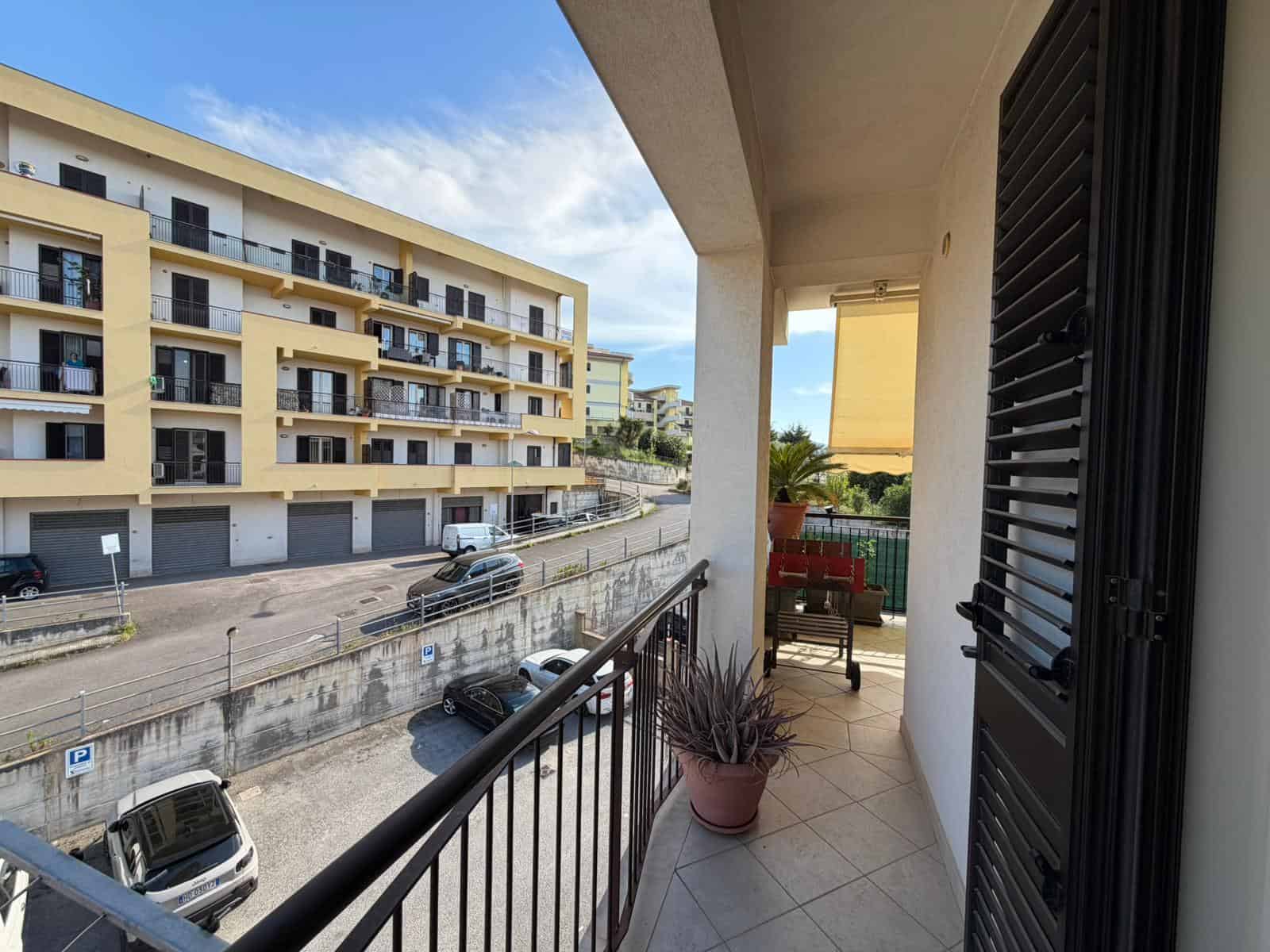Balcone di un appartamento con vista su edificio residenziale e parcheggio, piante decorative e sedie, in un ambiente luminoso e accogliente, ideale per relax e momenti all'aperto.