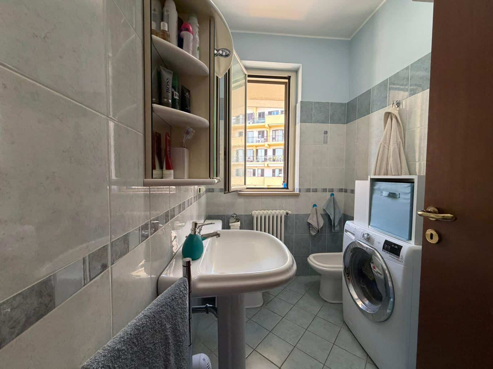 Lavatrice e lavabo in un bagno di piccole dimensioni con pareti rivestite di piastrelle beige e grigie, finestra che permette l'ingresso di luce naturale, e scaffale con prodotti per l'igiene personal.