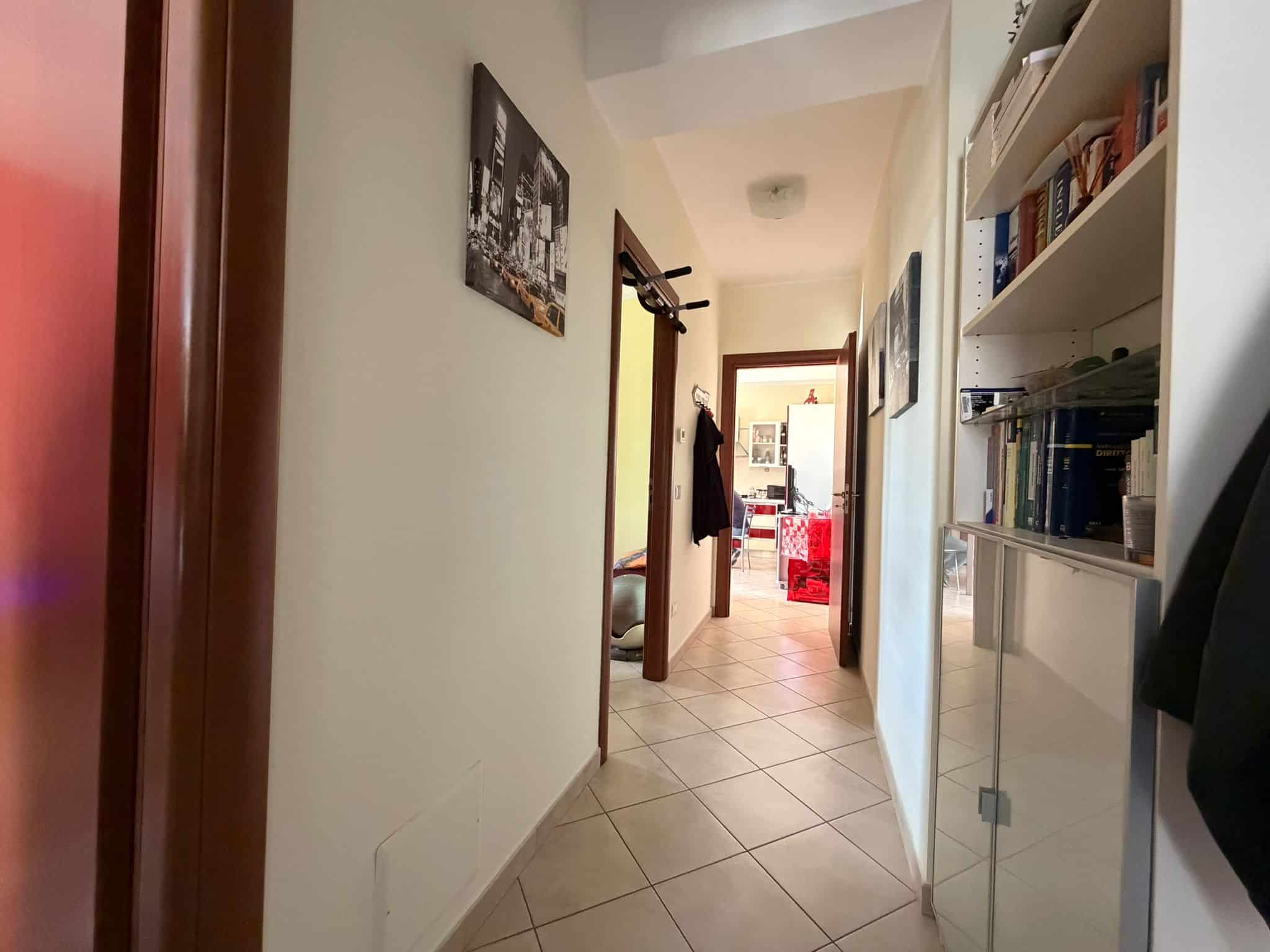 Ampia vista di un corridoio luminoso in un appartamento, con pareti chiare, scaffali con libri e decorazioni, che conduce a un soggiorno con divani rossi e grandi finestre, ideale per ambienti residen.