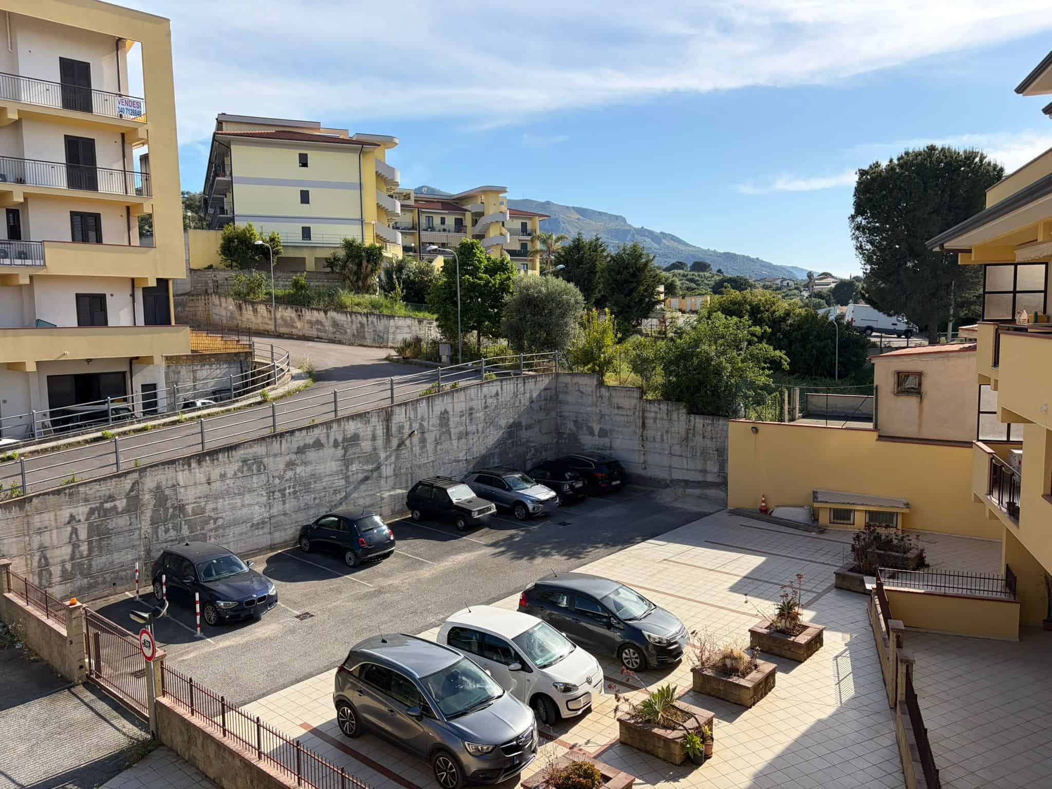 Auto in parcheggio all'aperto con edifici residenziali e montagne sullo sfondo, in una giornata soleggiata.