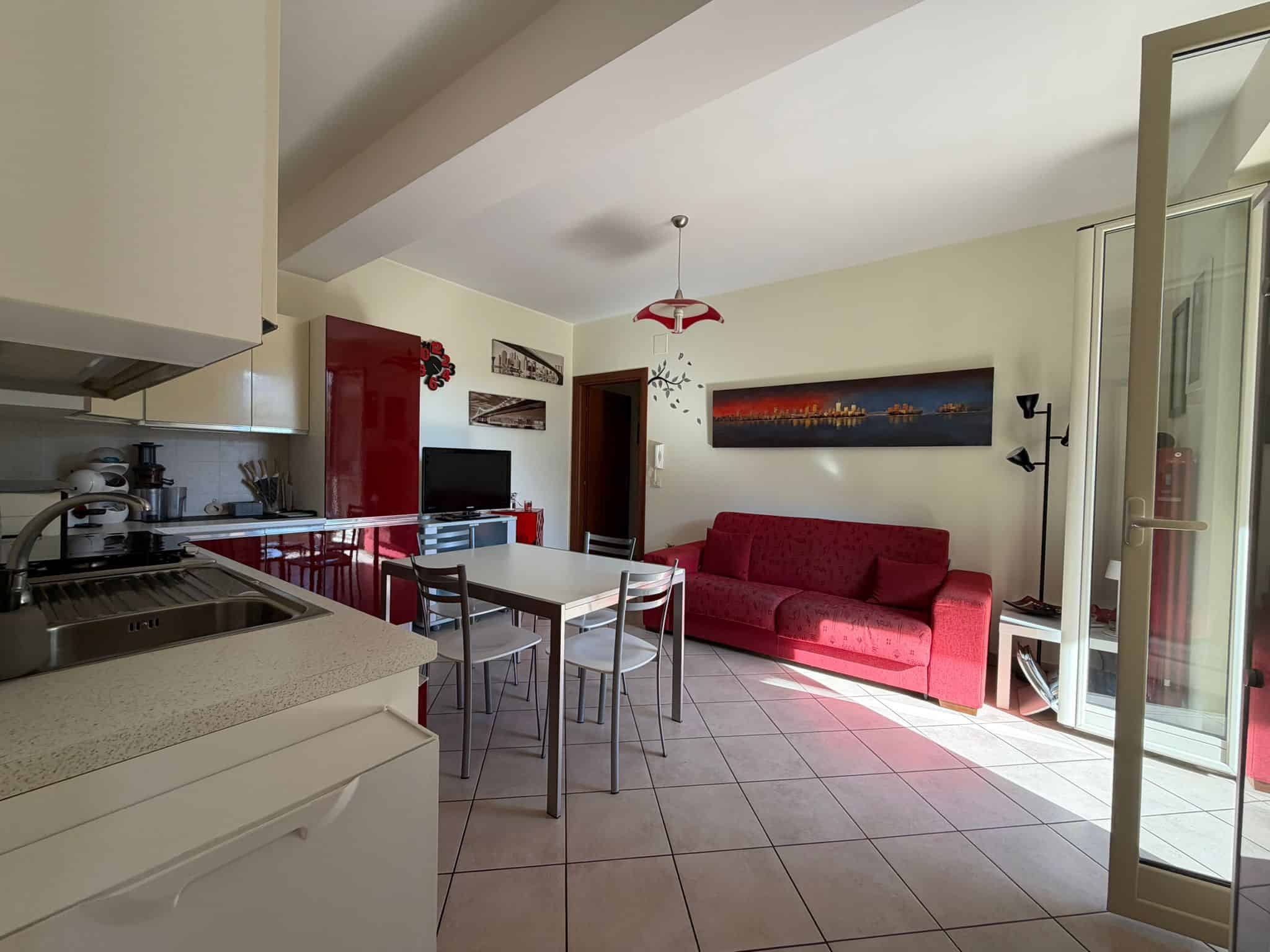 Interno di un soggiorno moderno con cucina aperta, divano rosso e accessori di design, illuminato dalla luce naturale proveniente dalla porta finestra.