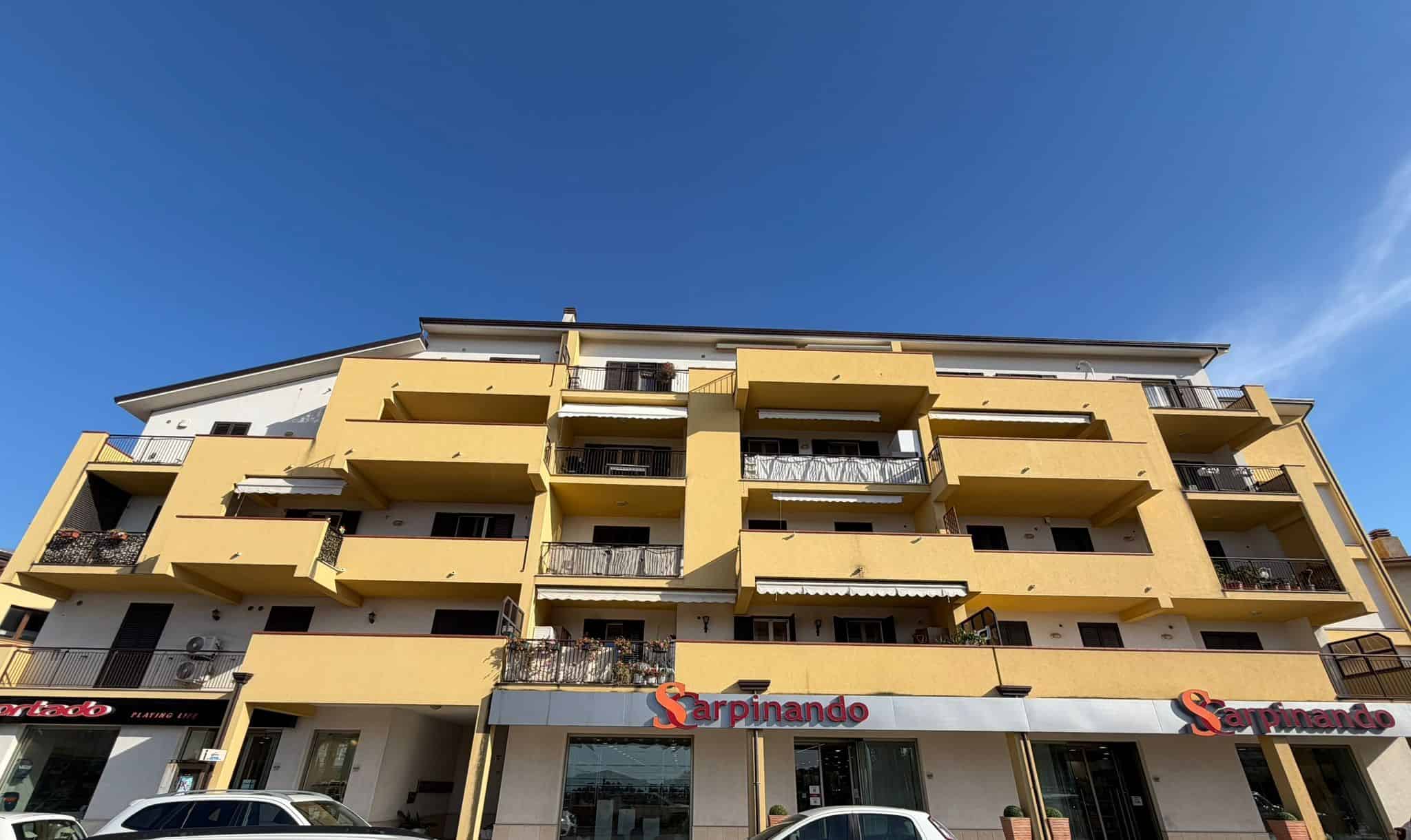 Ampio edificio residenziale con negozio al piano terra, facciata gialla e balconi spaziosi, situato in zona urbana ben servita, ideale per famiglie e investitori immobiliari.
