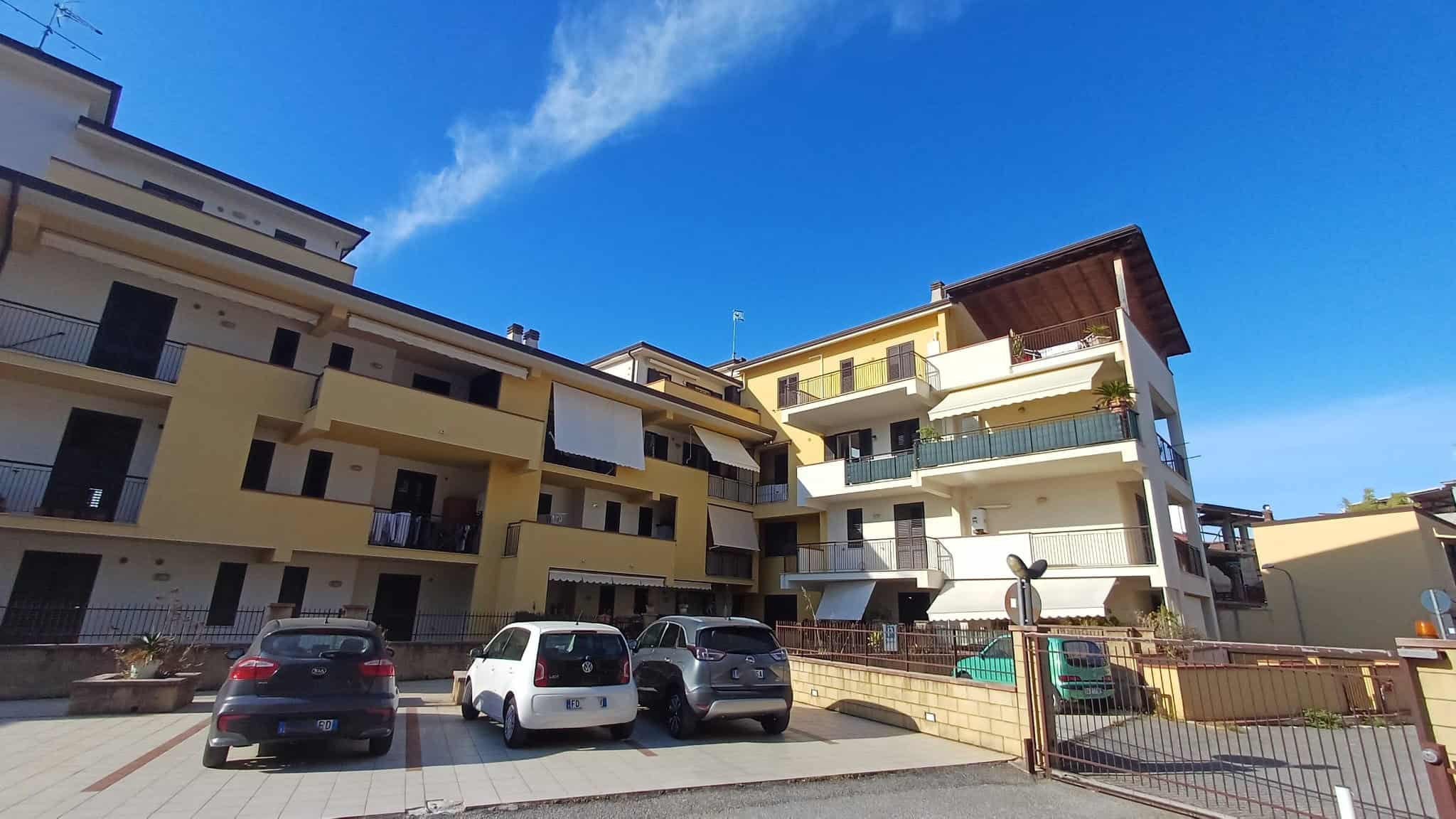 Parcheggio esterno di un complesso residenziale moderno con appartamenti, auto parcheggiate e balconi, sotto un cielo azzurro con poche nuvole, in un contesto urbano di alta qualità.