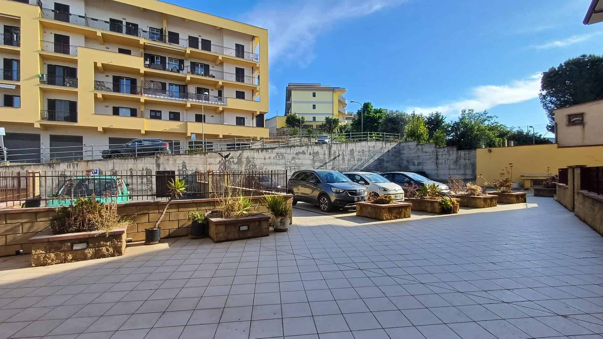 Panche di fiori e piante in un cortile urbano con parcheggio, sotto un cielo sereno, in un'area residenziale moderna. Ideale per immagini di spazi esterni e giardini in contesti immobiliari.