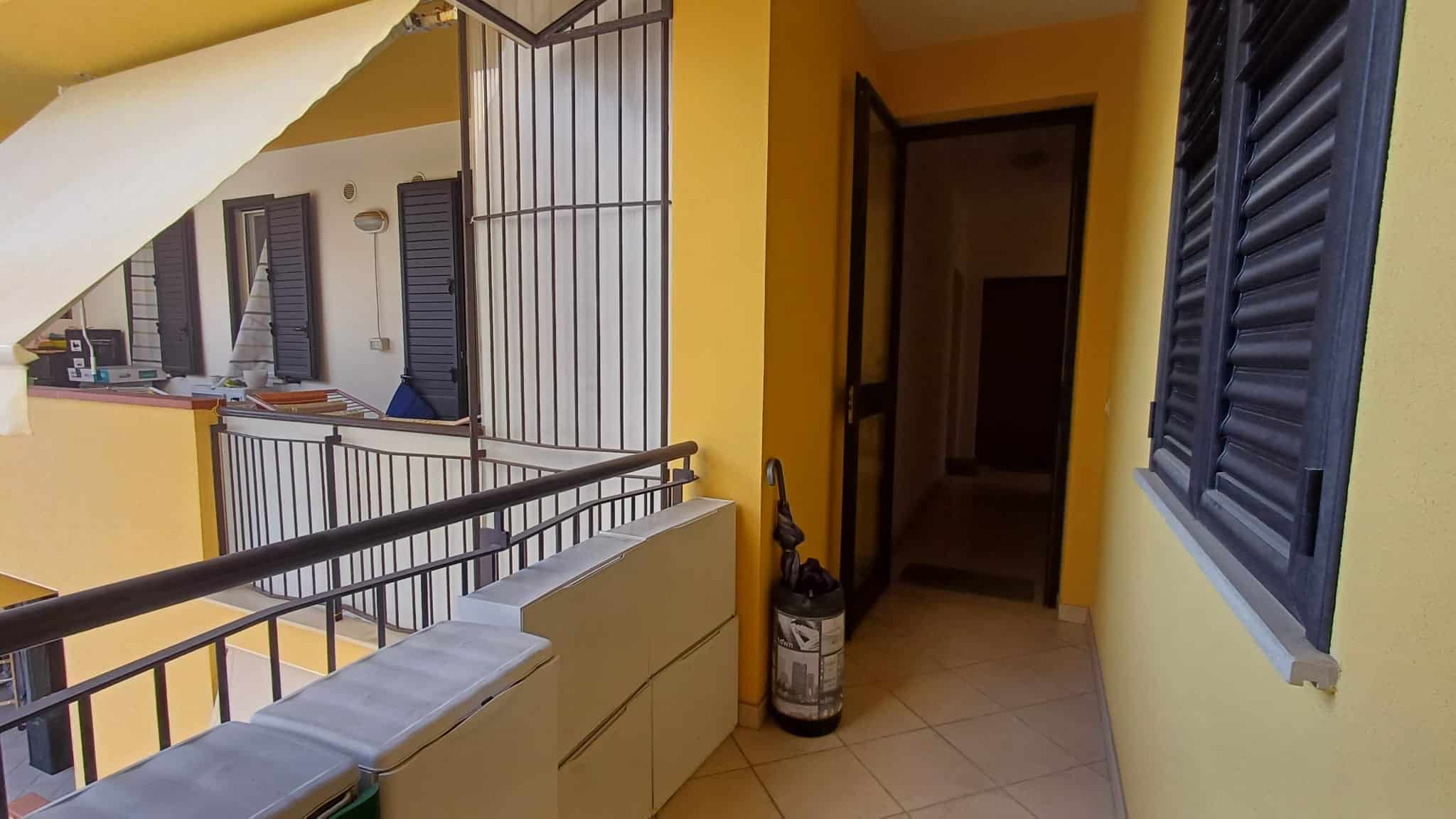 Balcone di un appartamento con ringhiera in metallo, pareti gialle e finestre con persiane blu, ambiente luminoso e accogliente, ideale per relax e momenti all'aperto.