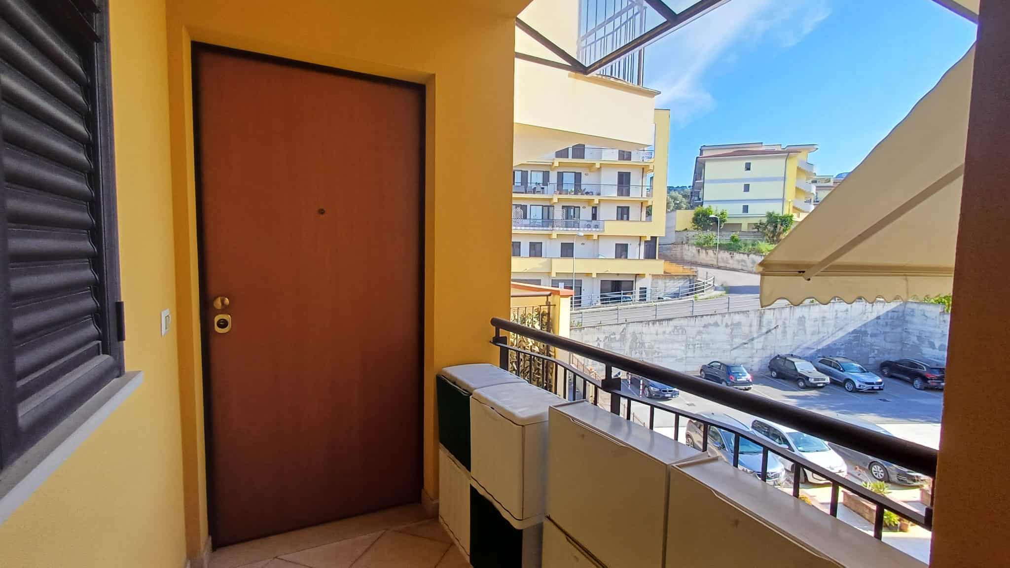 Porta d'ingresso gialla con balcone e vista su complesso residenziale moderno, illuminata dal sole, in un ambiente urbano e luminoso.